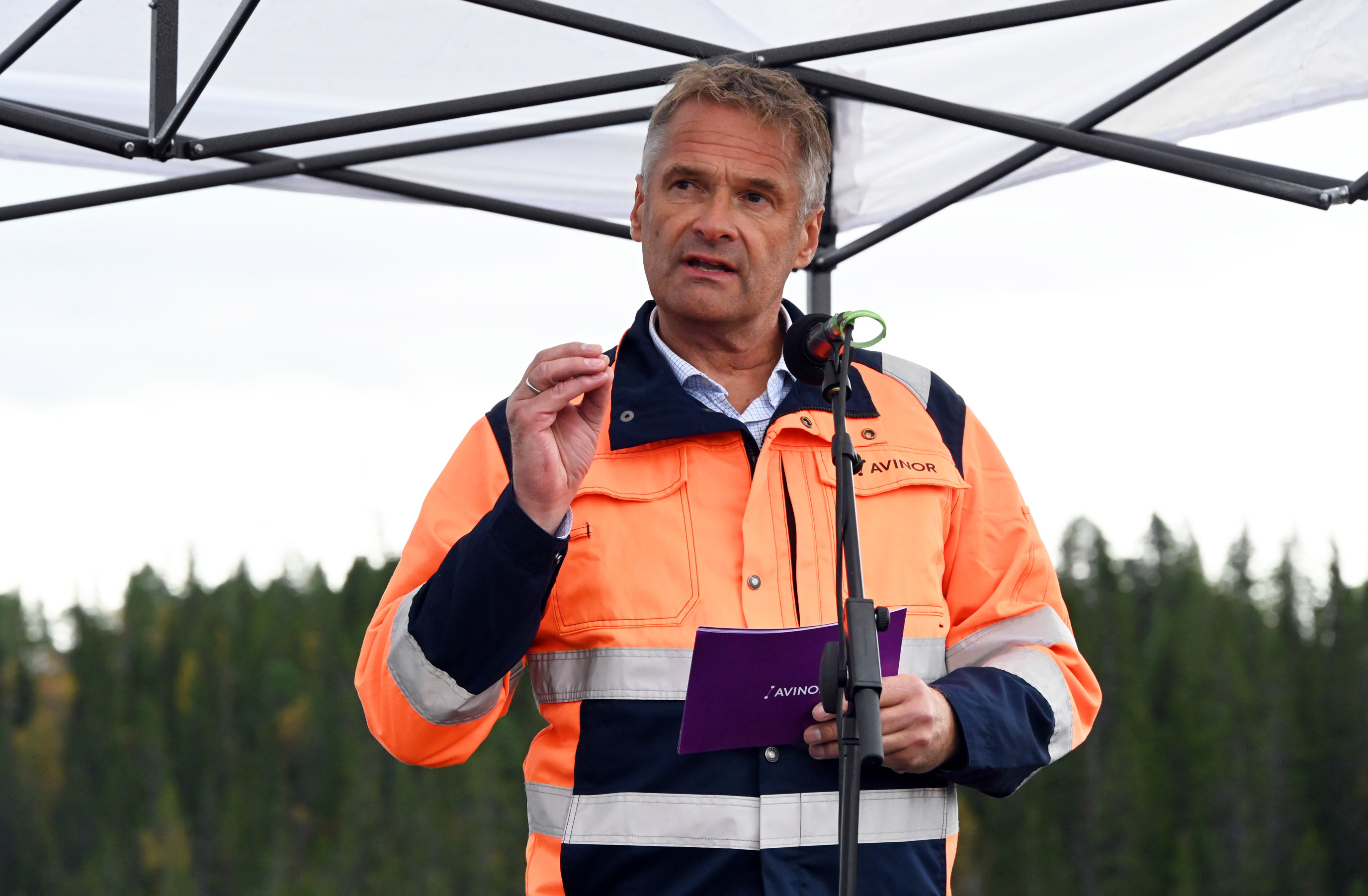 En mann i en oransje og blå Avinor-uniform holder en tale under en markise, med skog i bakgrunnen