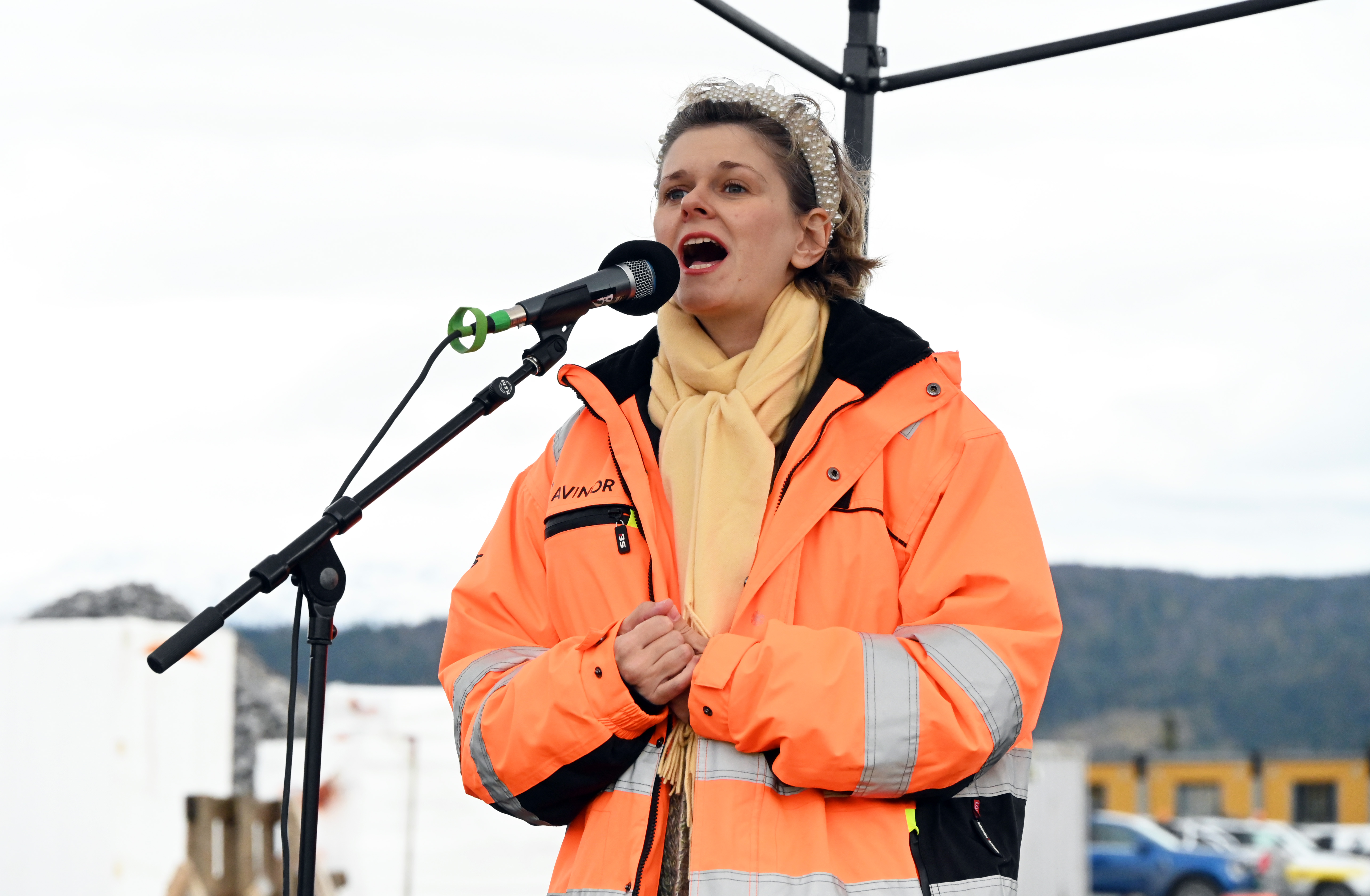 Kvinne i oransje refleksjakke synger i mikrofon under utendørs arrangement