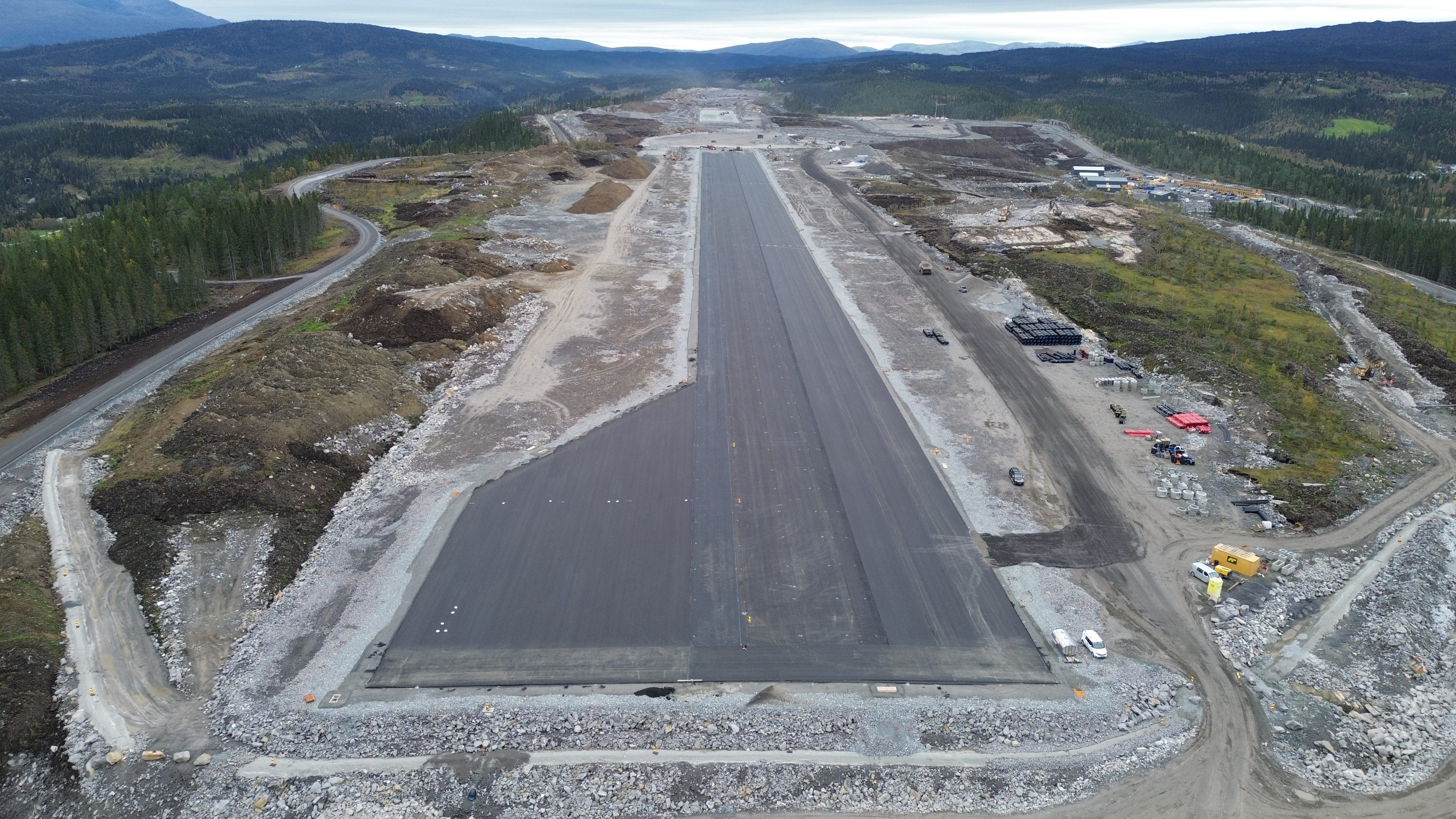 Flyfoto av en delvis konstruert rullebane omgitt av skog og fjellterreng, med veier og byggearbeid rundt
