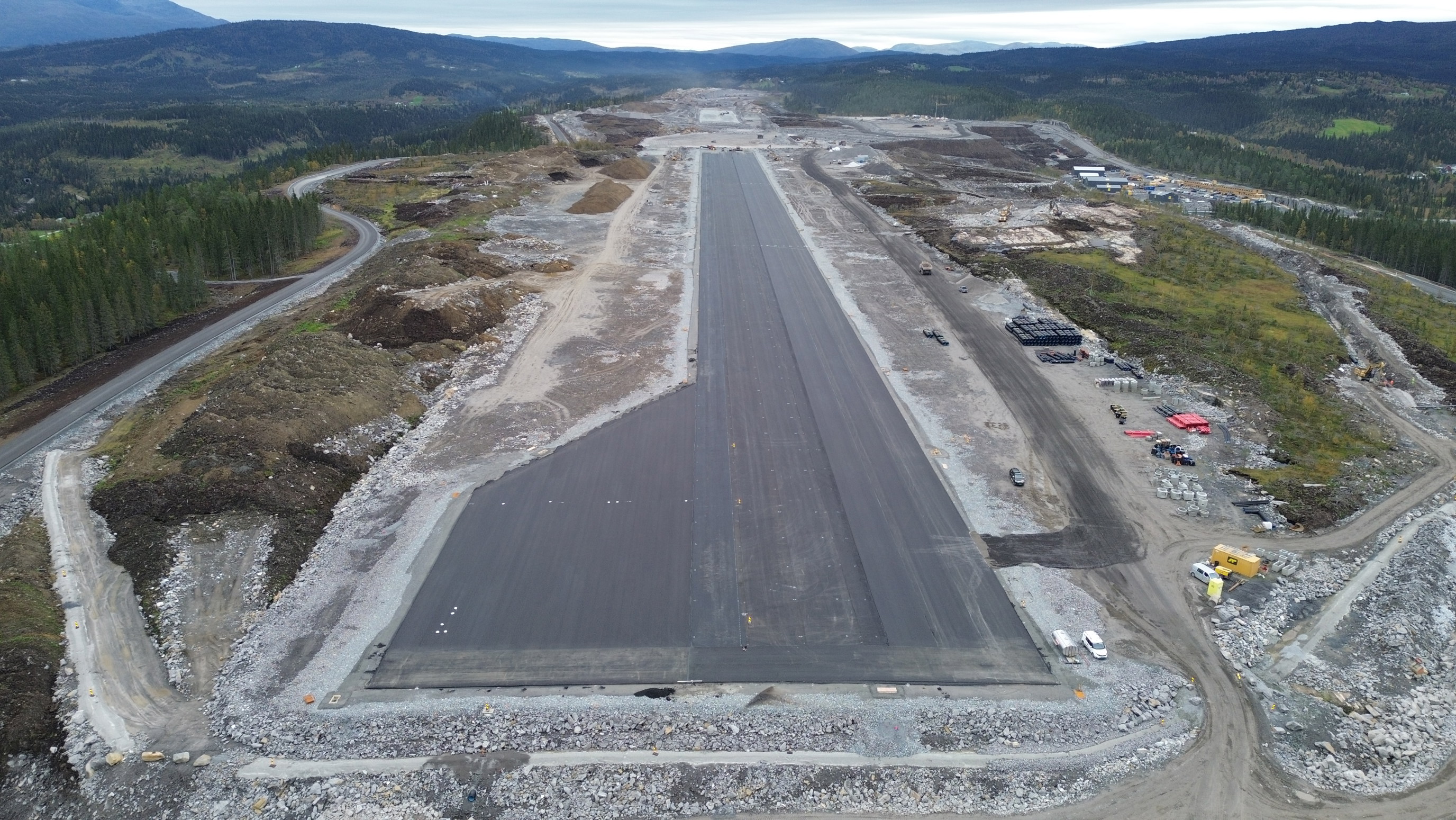 Flyfoto av en delvis konstruert rullebane omgitt av skog og fjellterreng, med veier og byggearbeid rundt