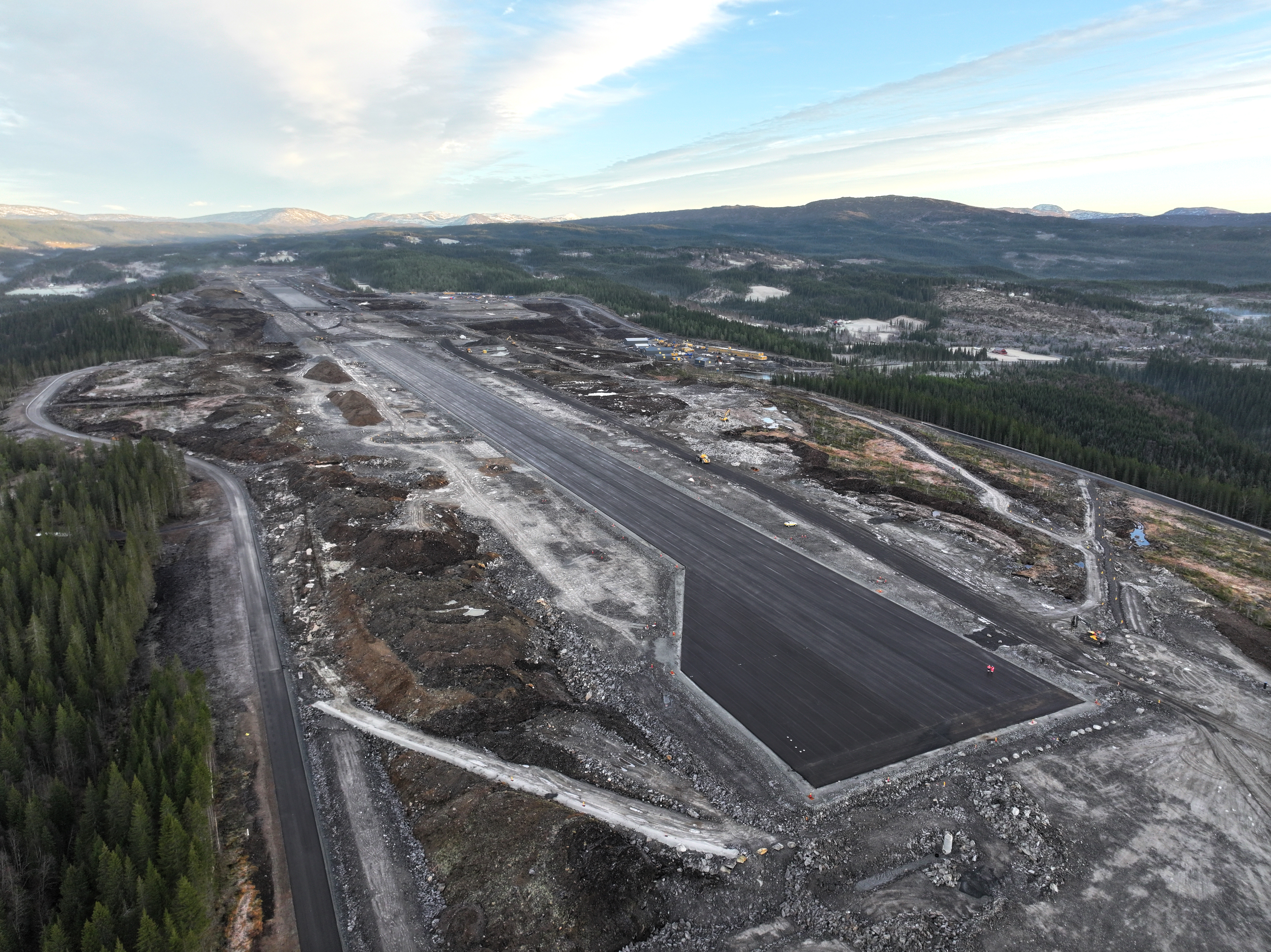 Luftfoto av en byggeplass for en fremtidig flystripe omgitt av skoglandskap og fjell i horisonten, under en delvis skyet himmel