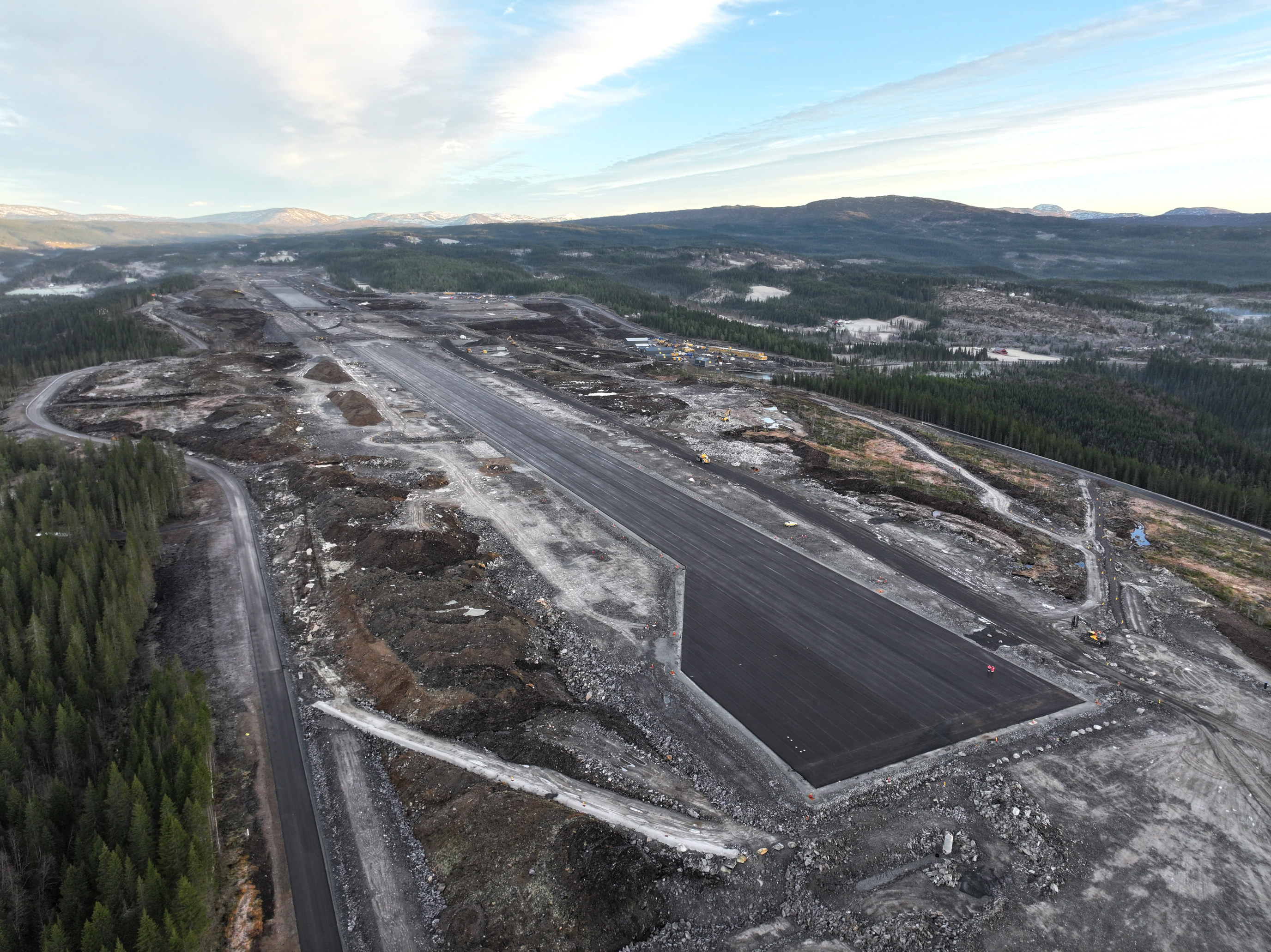 Luftfoto av en byggeplass for en fremtidig flystripe omgitt av skoglandskap og fjell i horisonten, under en delvis skyet himmel