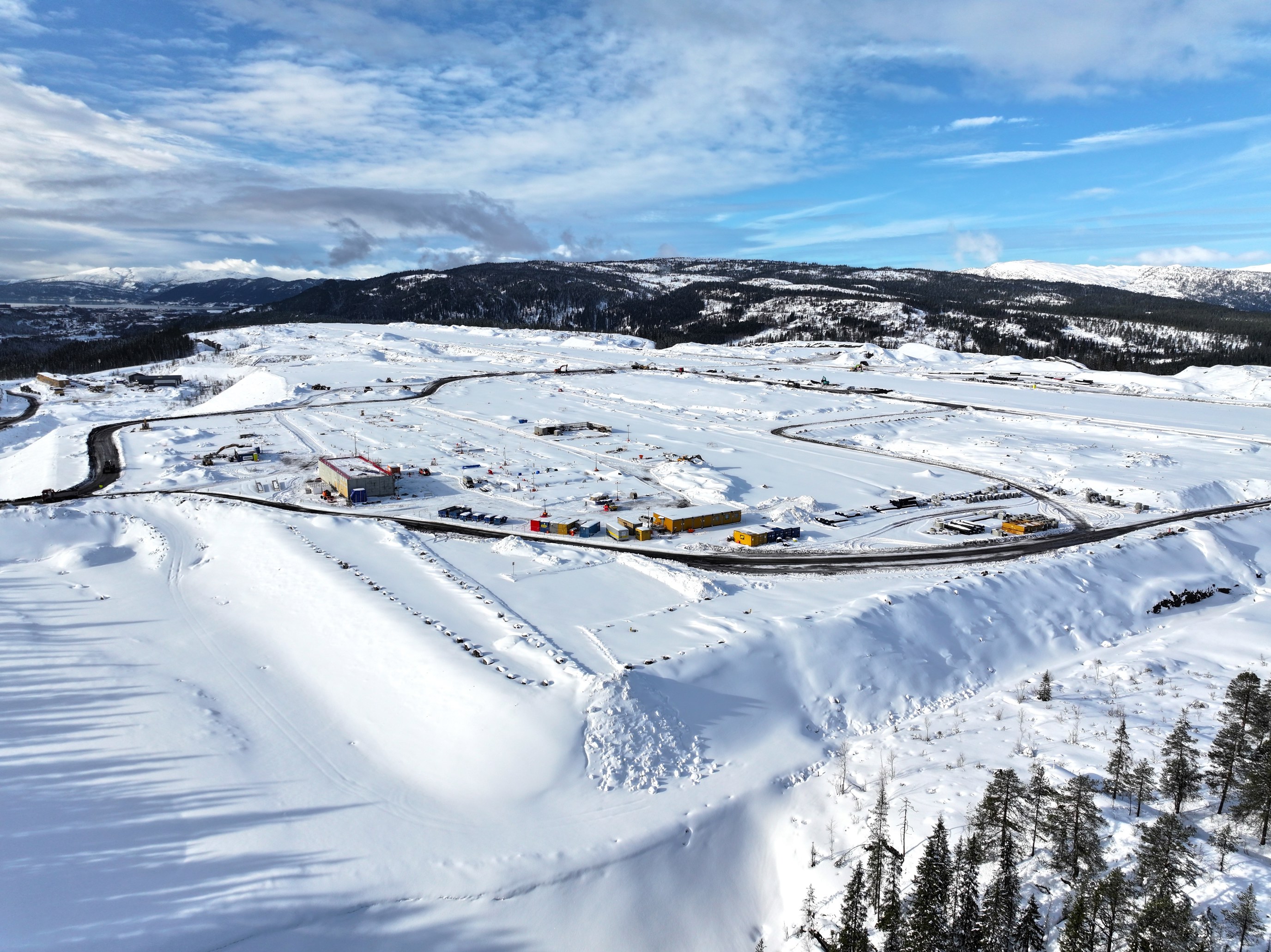 Flyfoto av et snødekt industriområde med flere bygninger og veier i et vinterlandskap i Norge, omgitt av skogkledde åser under en blå himmel