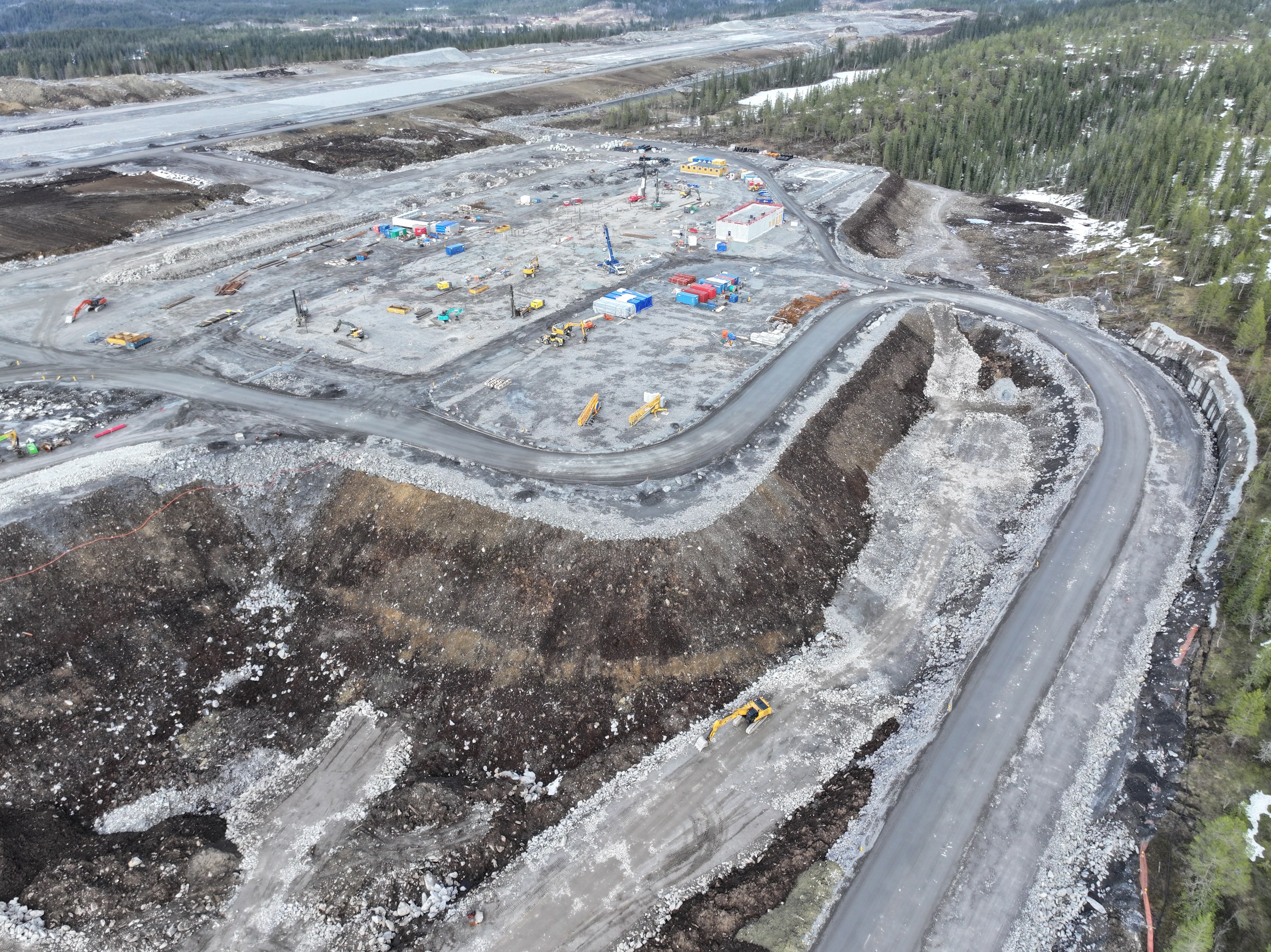 Luftfoto av en stor byggeplass med anleggsmaskiner, flere kjøretøy og bygninger i ulike byggefaser. Området er omgitt av skogslandskap med en svingete asfaltert vei langs kanten av anleggsområdet