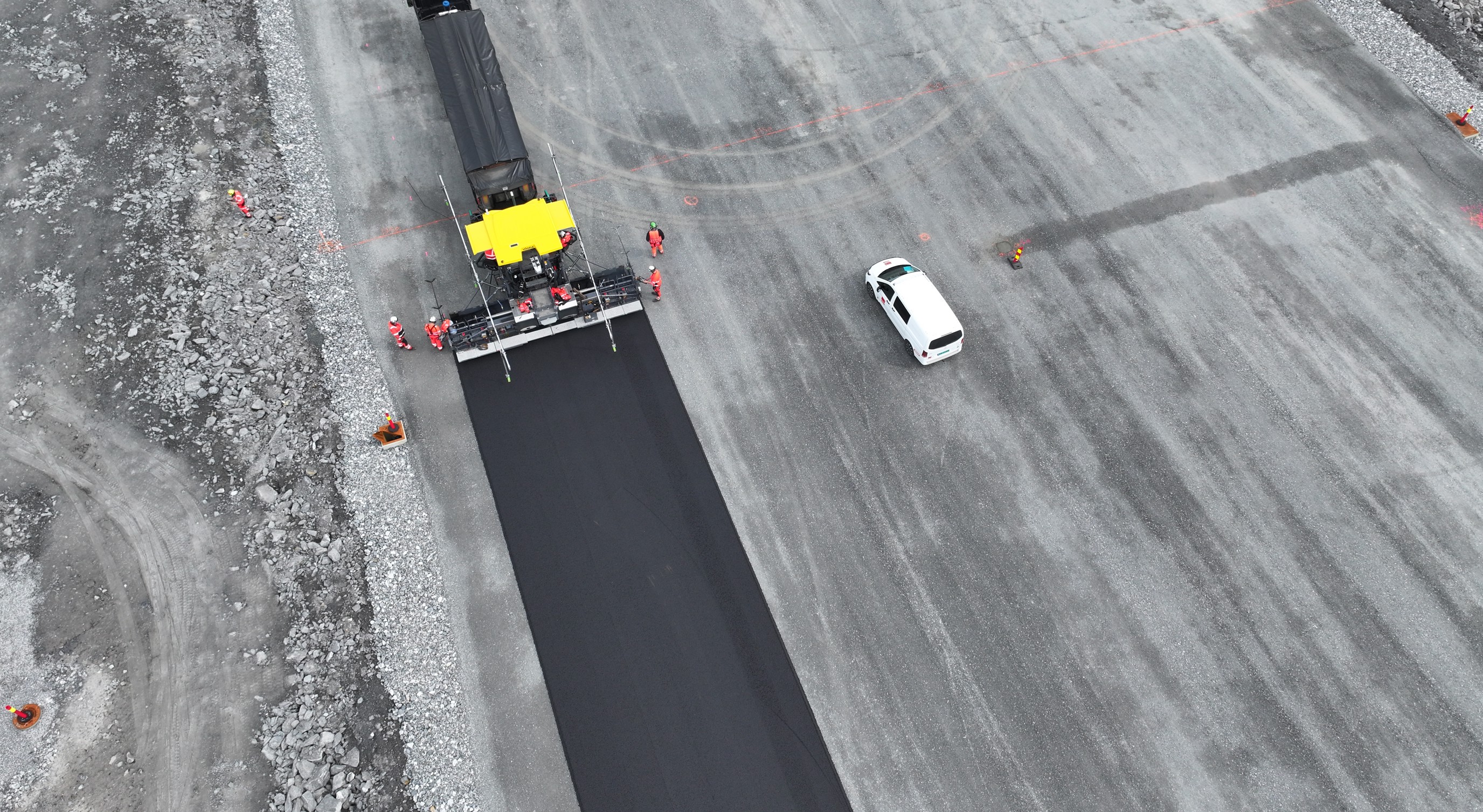 Luftfoto av en veiarbeidsplass hvor en asfaltlegger legger fersk asfalt på en bred vei. Flere arbeidere med oransje refleksklær står rundt maskinen, og en hvit bil står parkert ved siden av det nylagte asfaltfeltet. Området er omkranset av grus og steiner