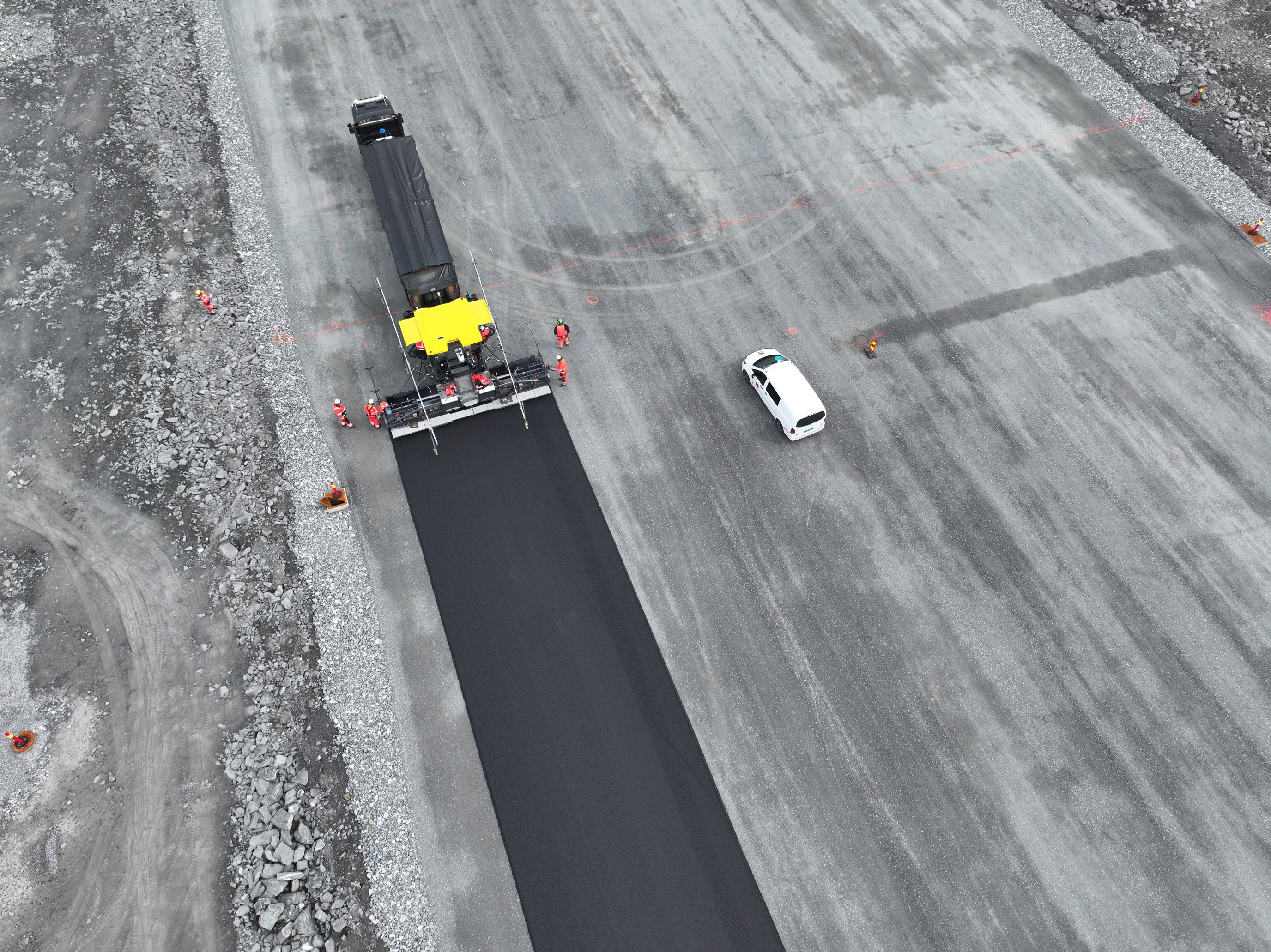 Luftfoto av en veiarbeidsplass hvor en asfaltlegger legger fersk asfalt på en bred vei. Flere arbeidere med oransje refleksklær står rundt maskinen, og en hvit bil står parkert ved siden av det nylagte asfaltfeltet. Området er omkranset av grus og steiner