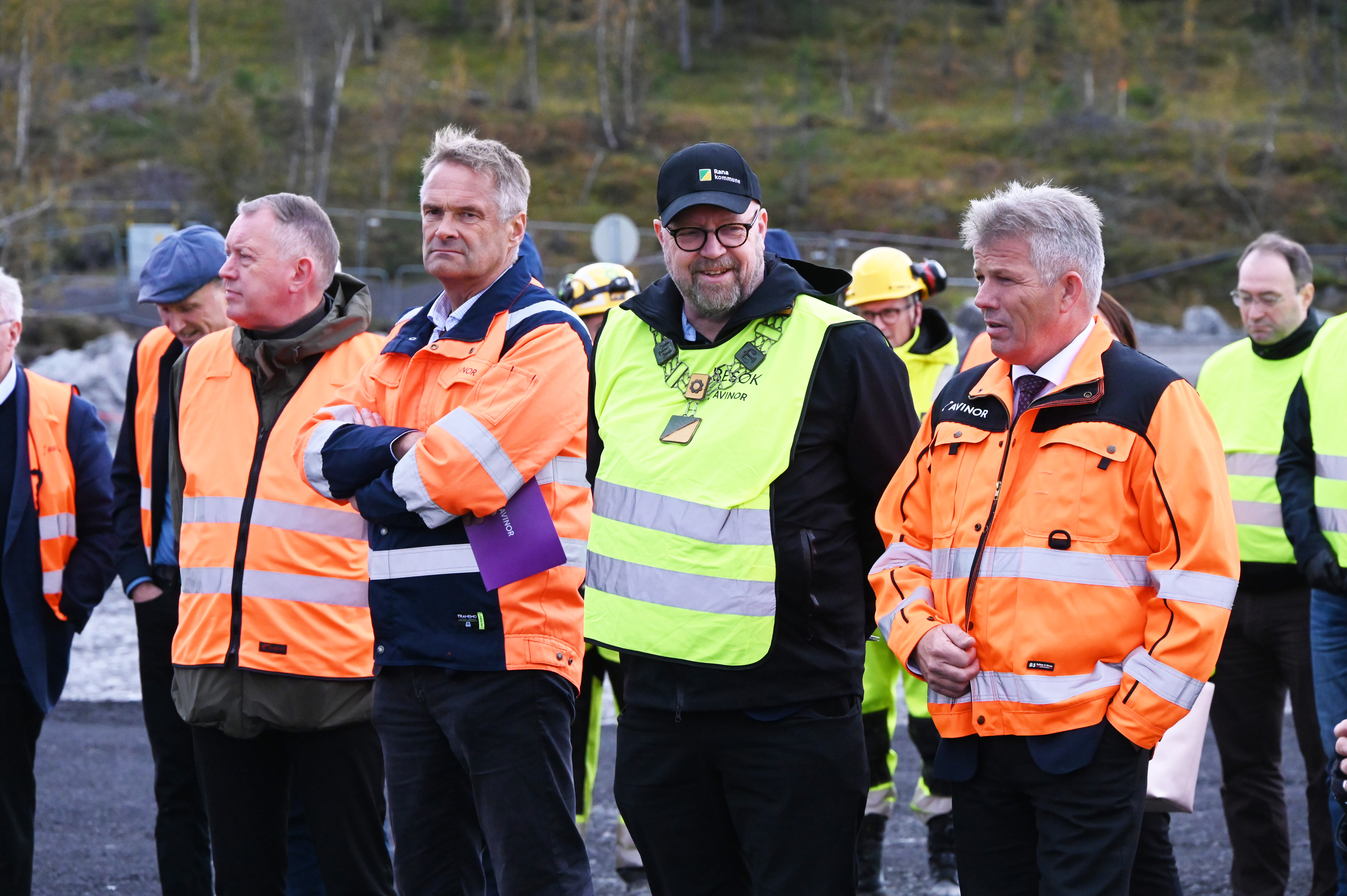 Personer i vernejakker og hjelmer på en byggeplass, samlet til gjennomgang av prosjektet. Omkringliggende natur med trær i bakgrunnen