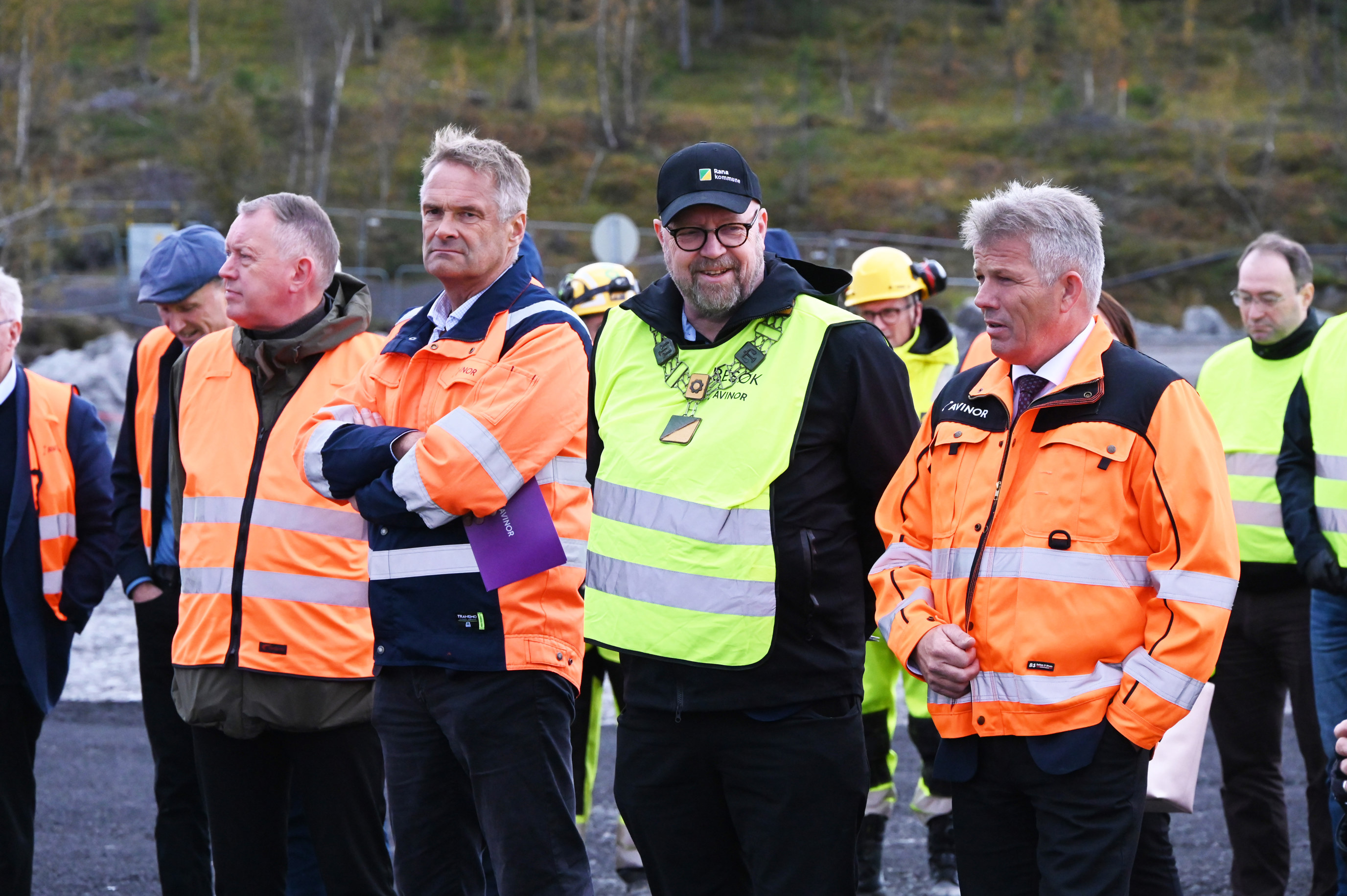 Personer i vernejakker og hjelmer på en byggeplass, samlet til gjennomgang av prosjektet. Omkringliggende natur med trær i bakgrunnen