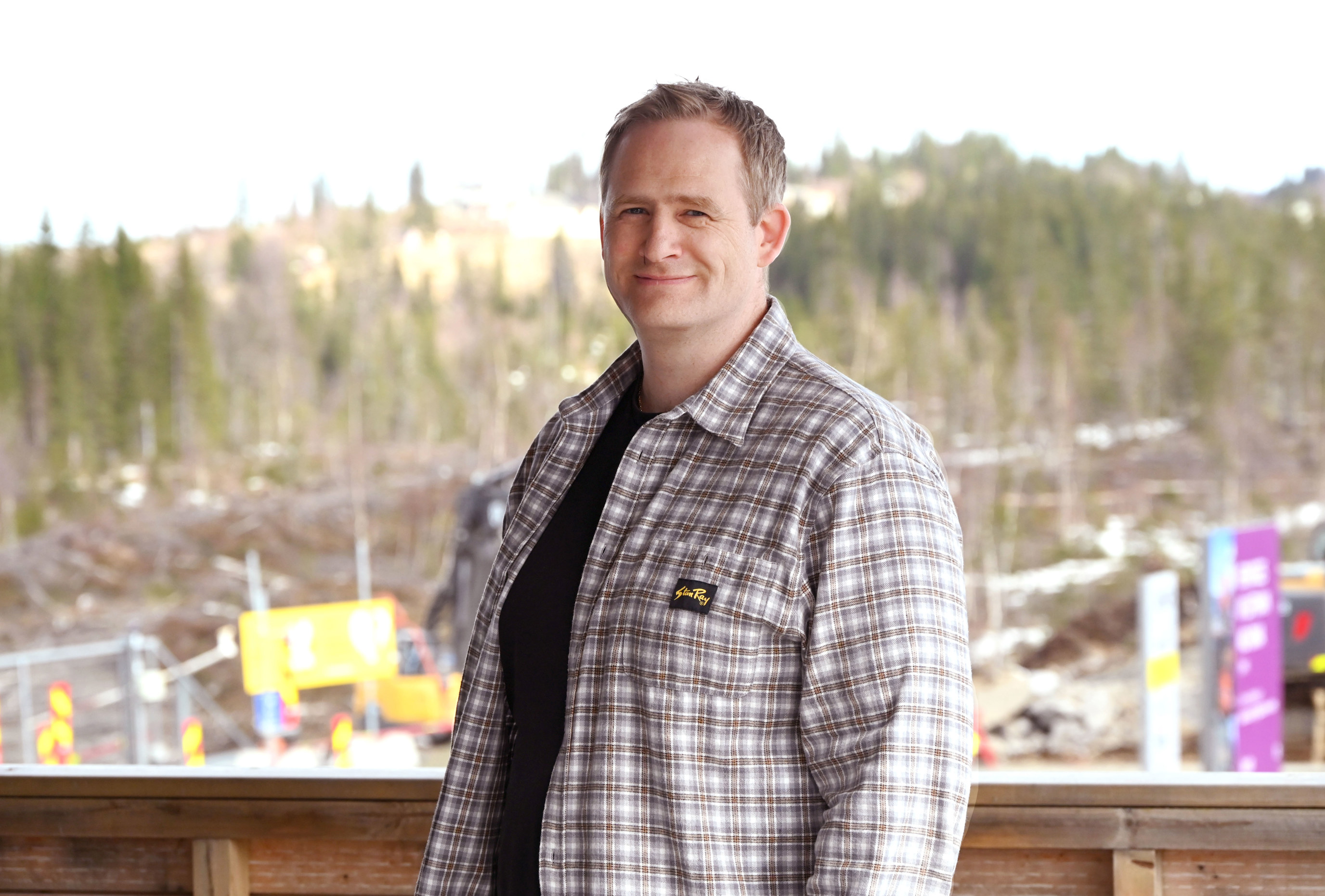 En mann med rutete skjorte står utendørs foran et skoglandskap med trær i bakgrunnen