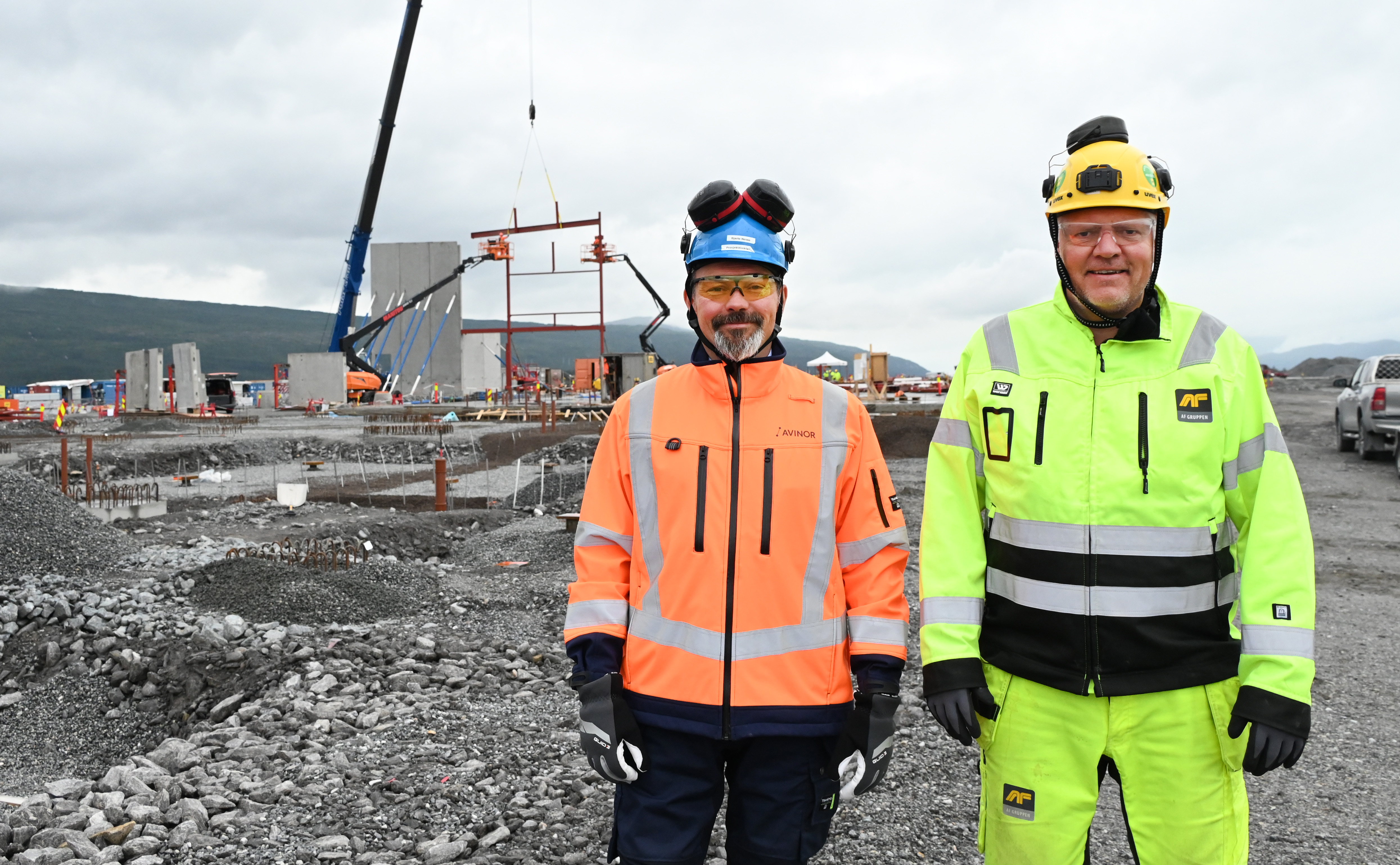 To bygningsarbeidere i verneutstyr står på en byggeplass med kran og betongvegger i bakgrunnen
