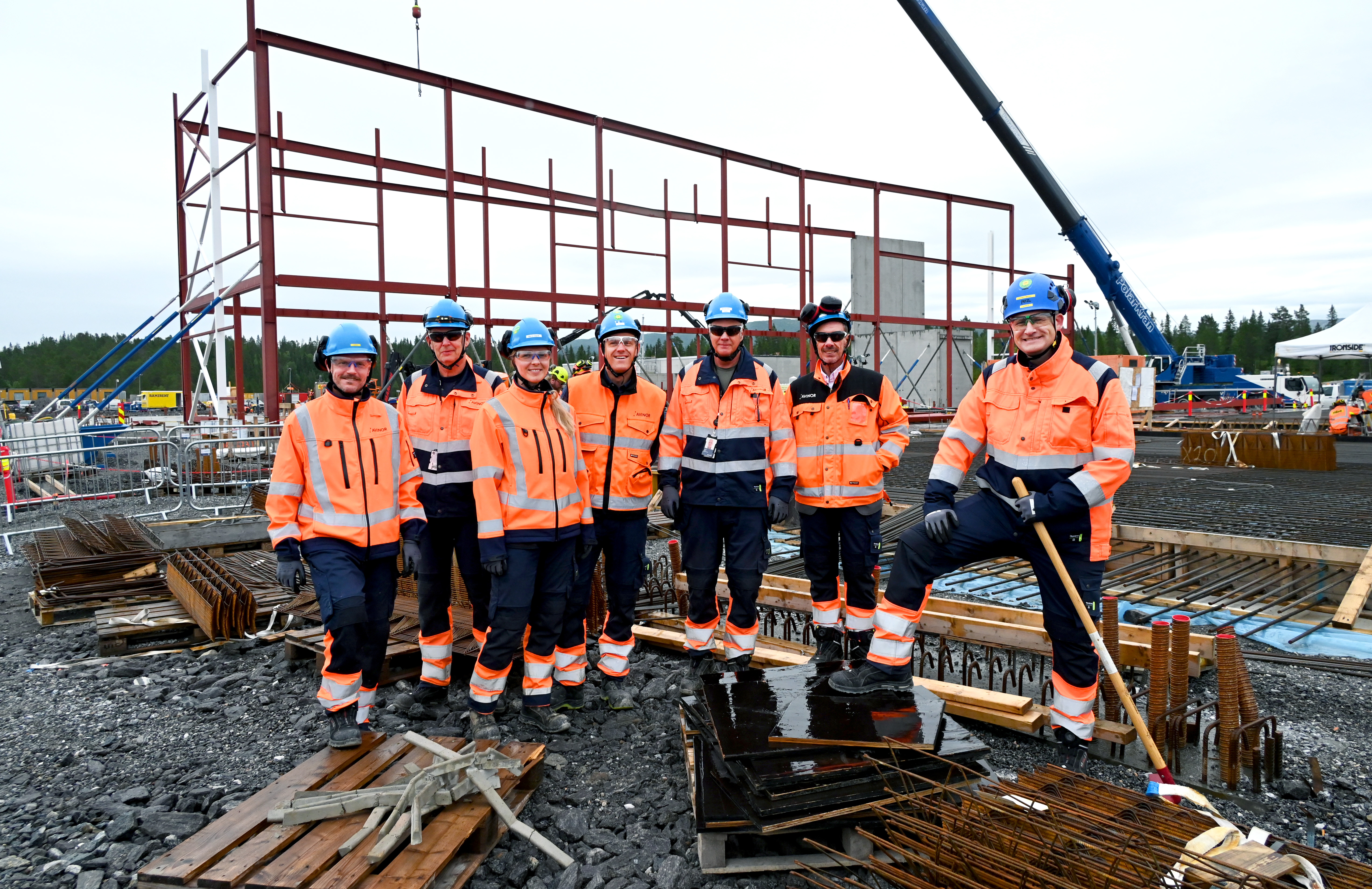 Bygningsarbeidere i verneutstyr på byggeplass, stålramme i bakgrunnen, kran og skog