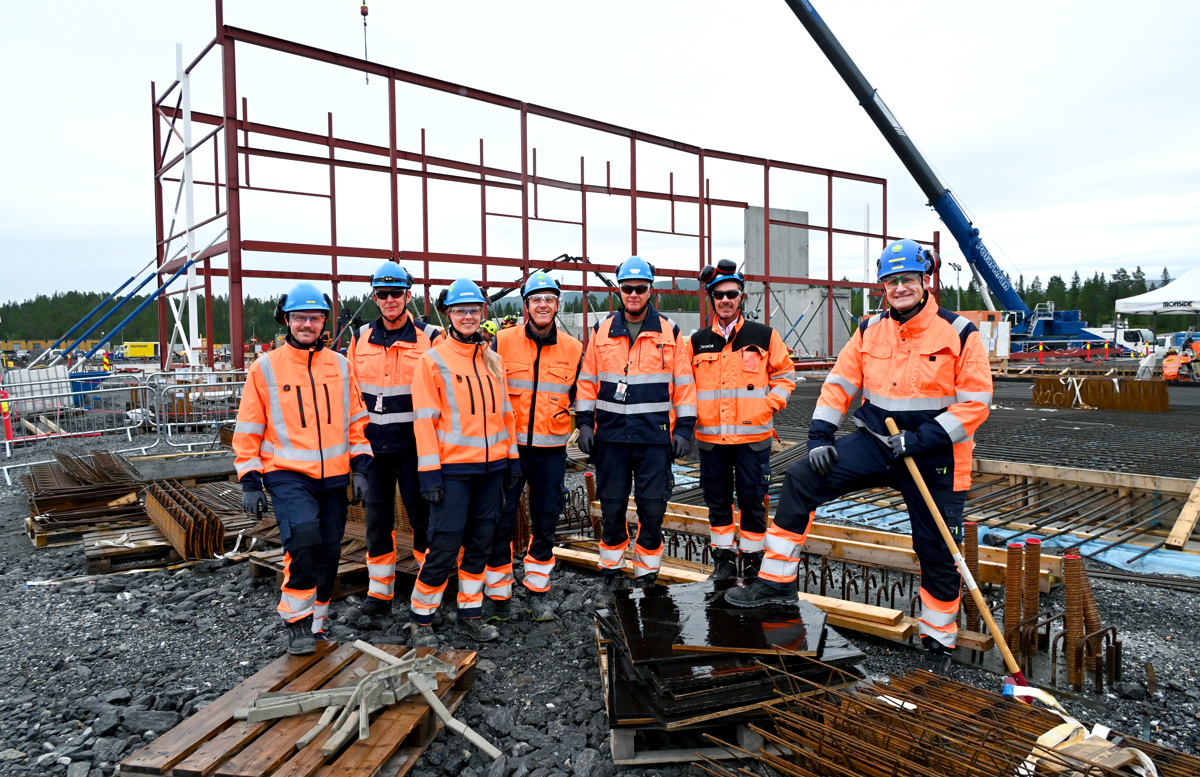 Bygningsarbeidere i verneutstyr på byggeplass, stålramme i bakgrunnen, kran og skog