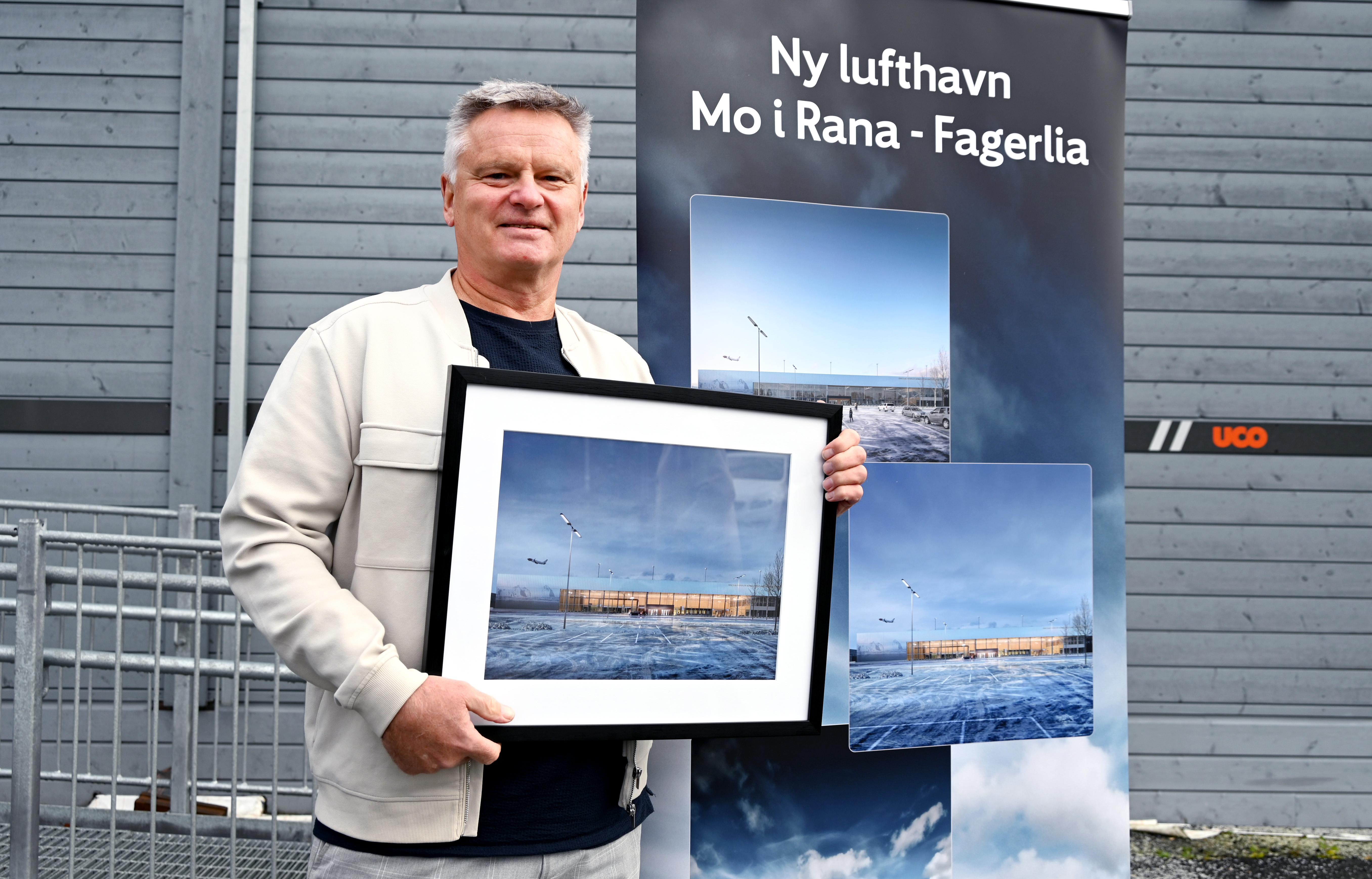 Person holder et innrammet bilde foran en plakat som annonserer 