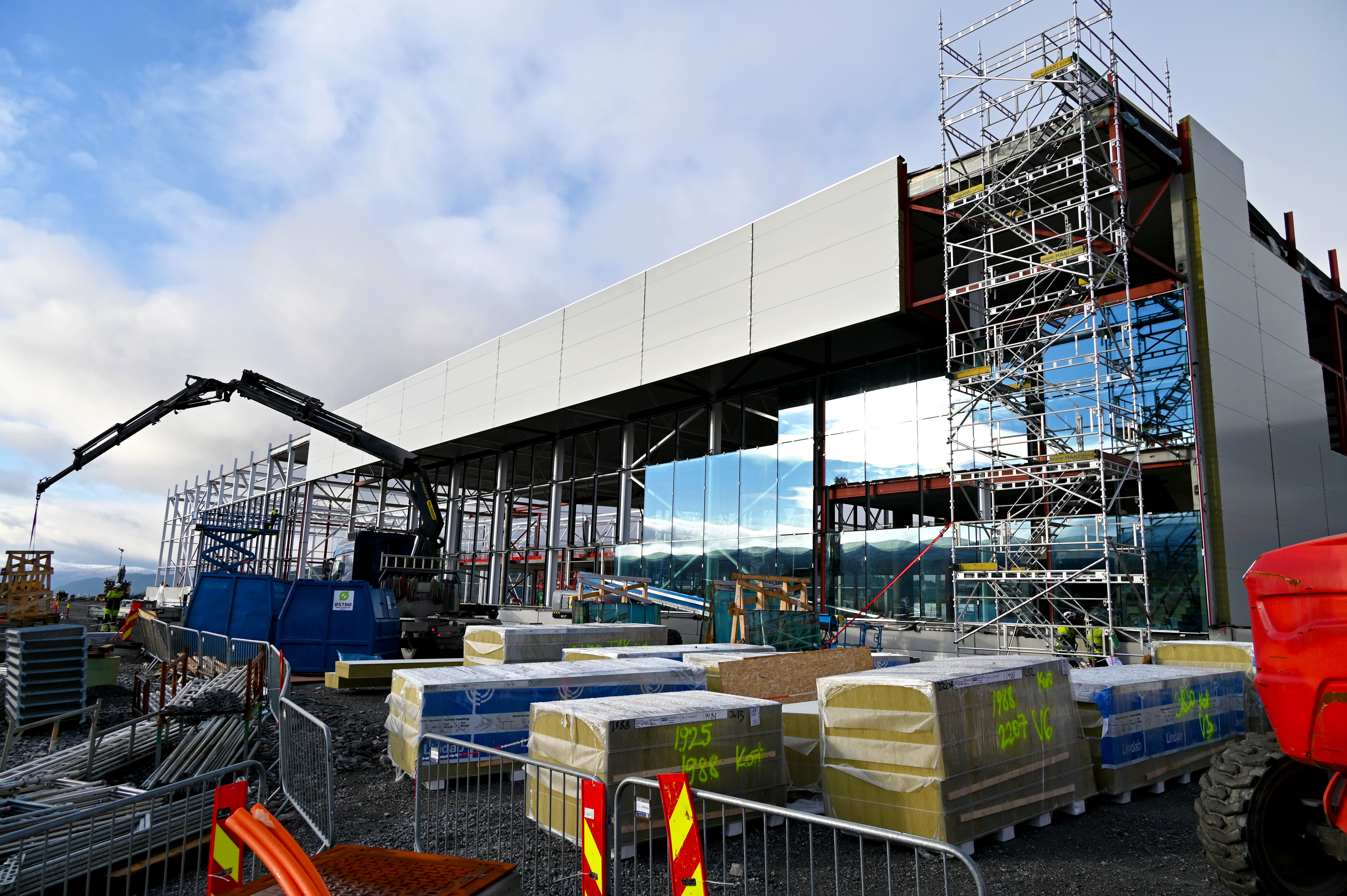 Byggeplass med konstruksjon av moderne industribygg, hvor arbeidere bruker stillaser og tungt maskineri. Reflekterende glassfasade installeres, omgitt av byggematerialer