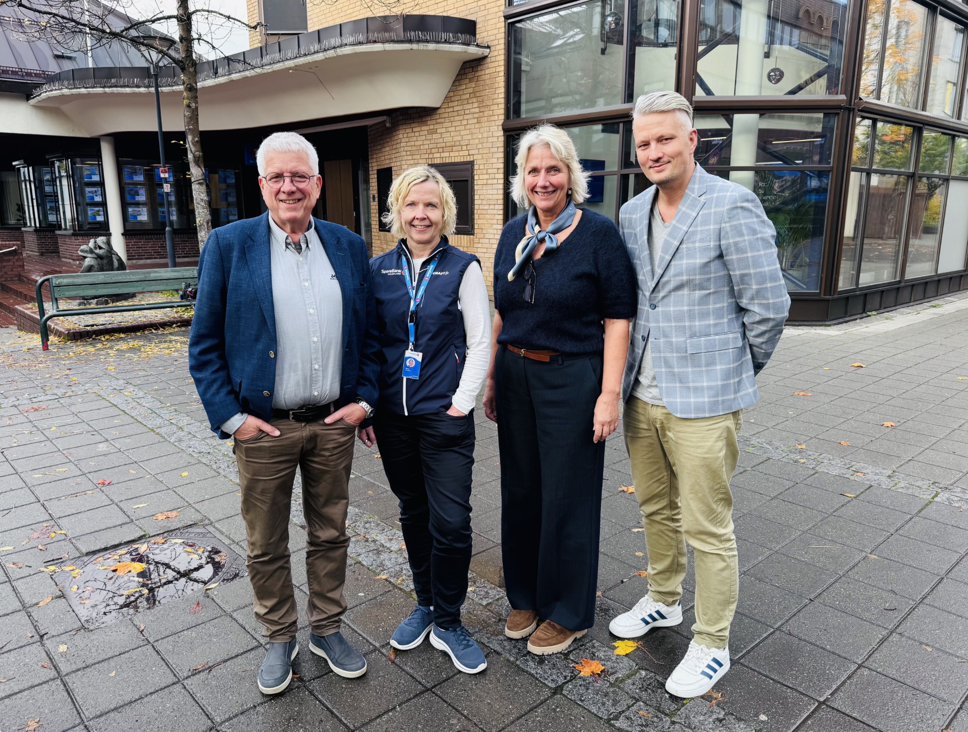 Fire personer står ute foran en murbygning i høstvær, alle kledd i casual antrekk med smilende ansikter