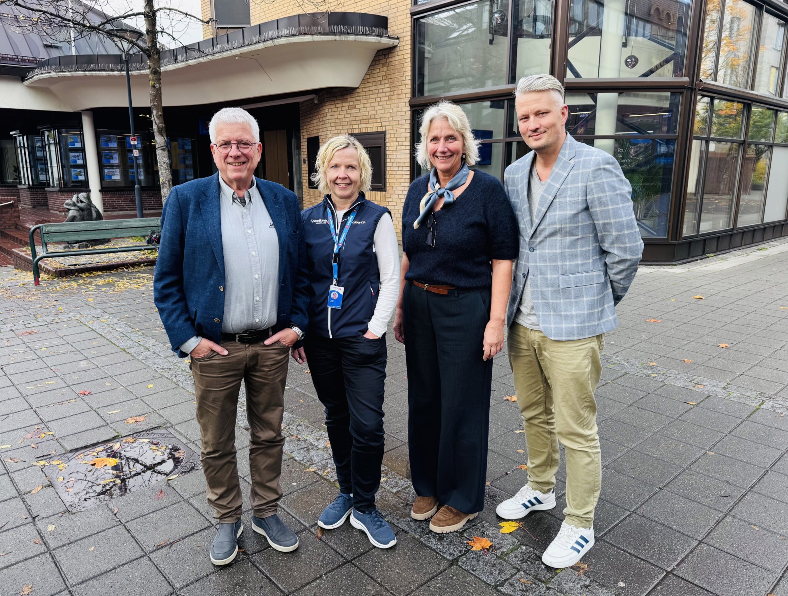 Fire personer står ute foran en murbygning i høstvær, alle kledd i casual antrekk med smilende ansikter