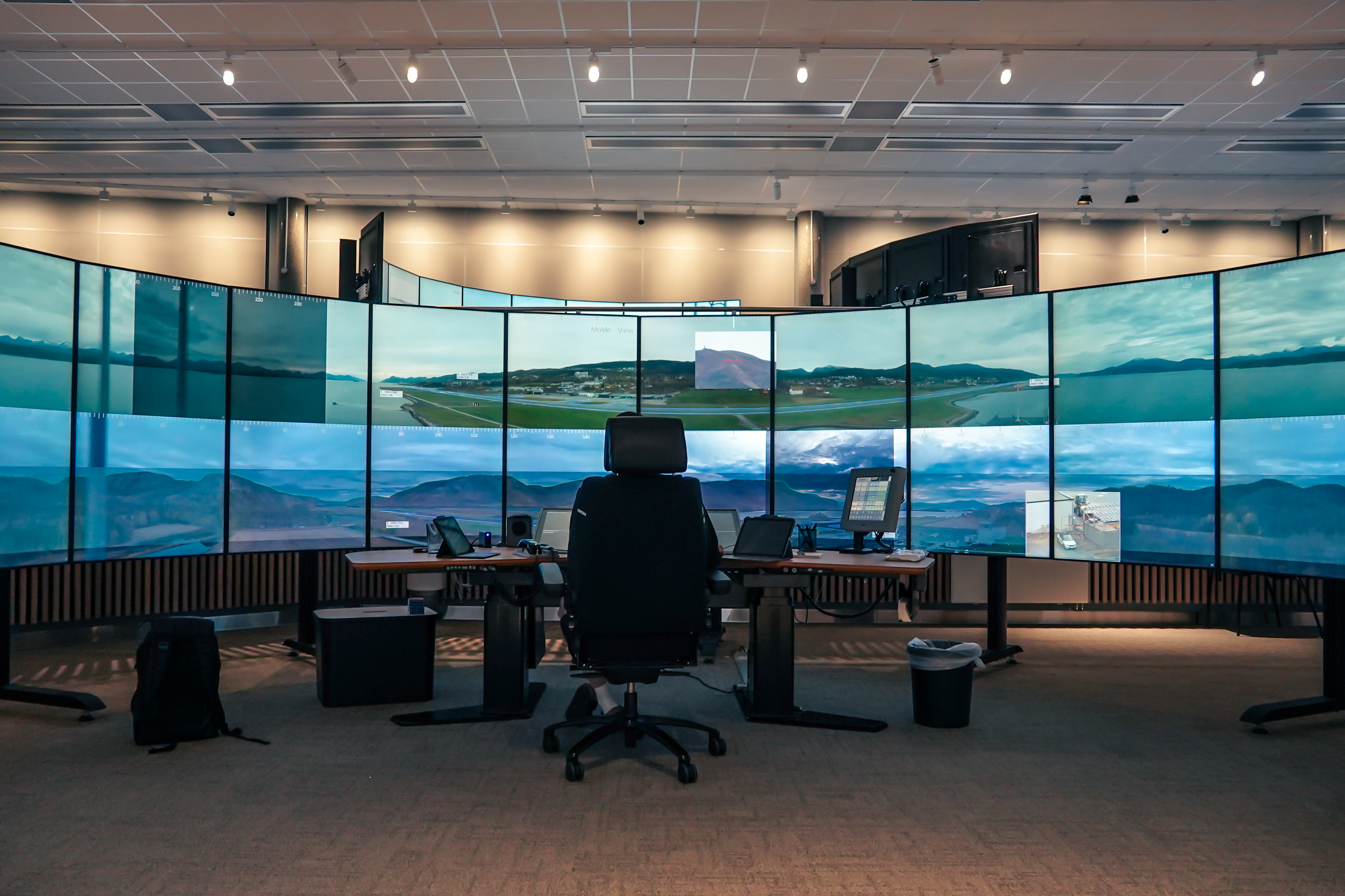 Stor luftfartskontrollrom med flere skjermer som viser flyplasser og luftutsikt. Sentral pult med stol, skjermer og utstyr under taklys i et høyteknologisk miljø