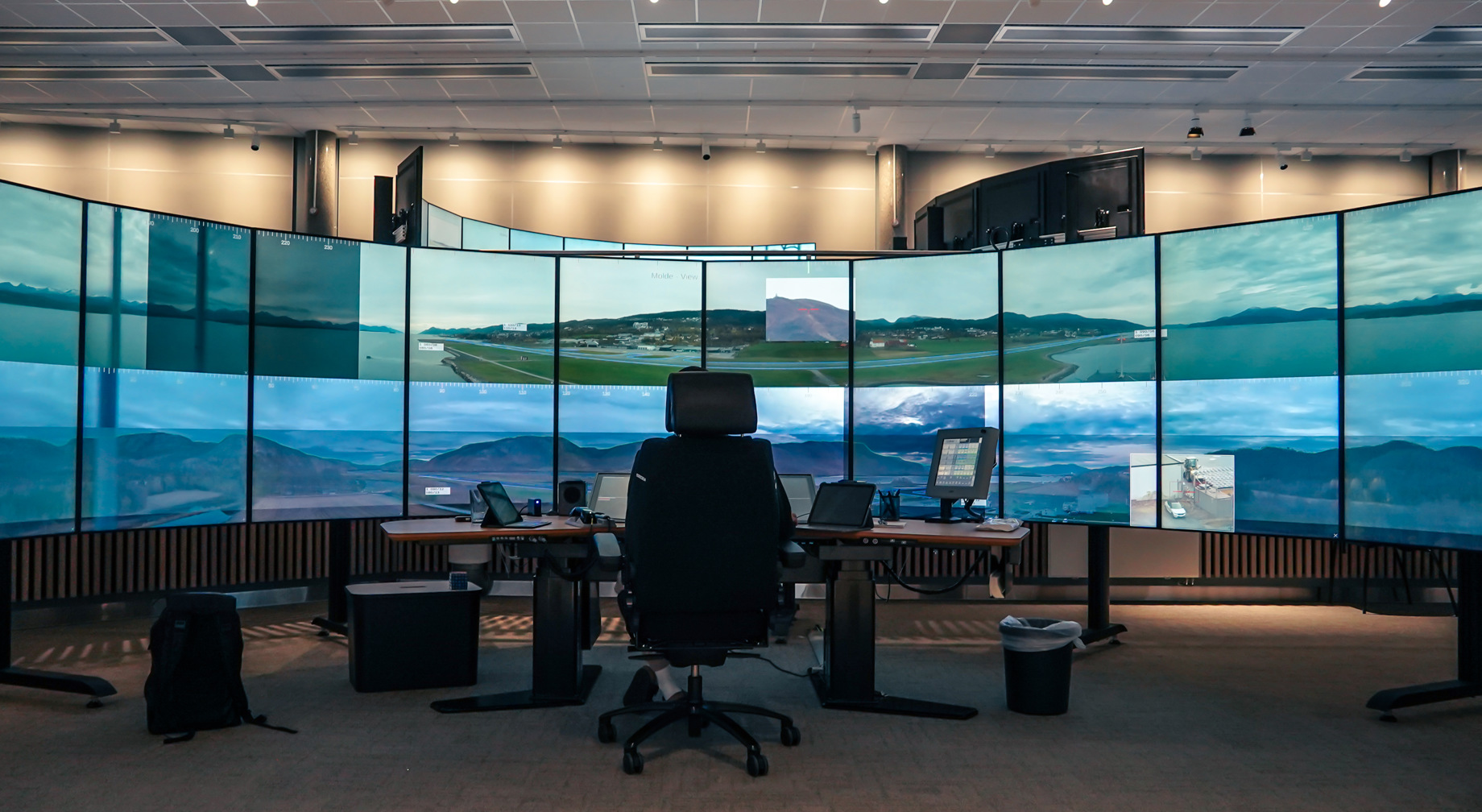 Ein kontrollrom med fleire store skjermer som viser landskap og flyplassområde, med ein stol og skrivebord framfor skjermane
