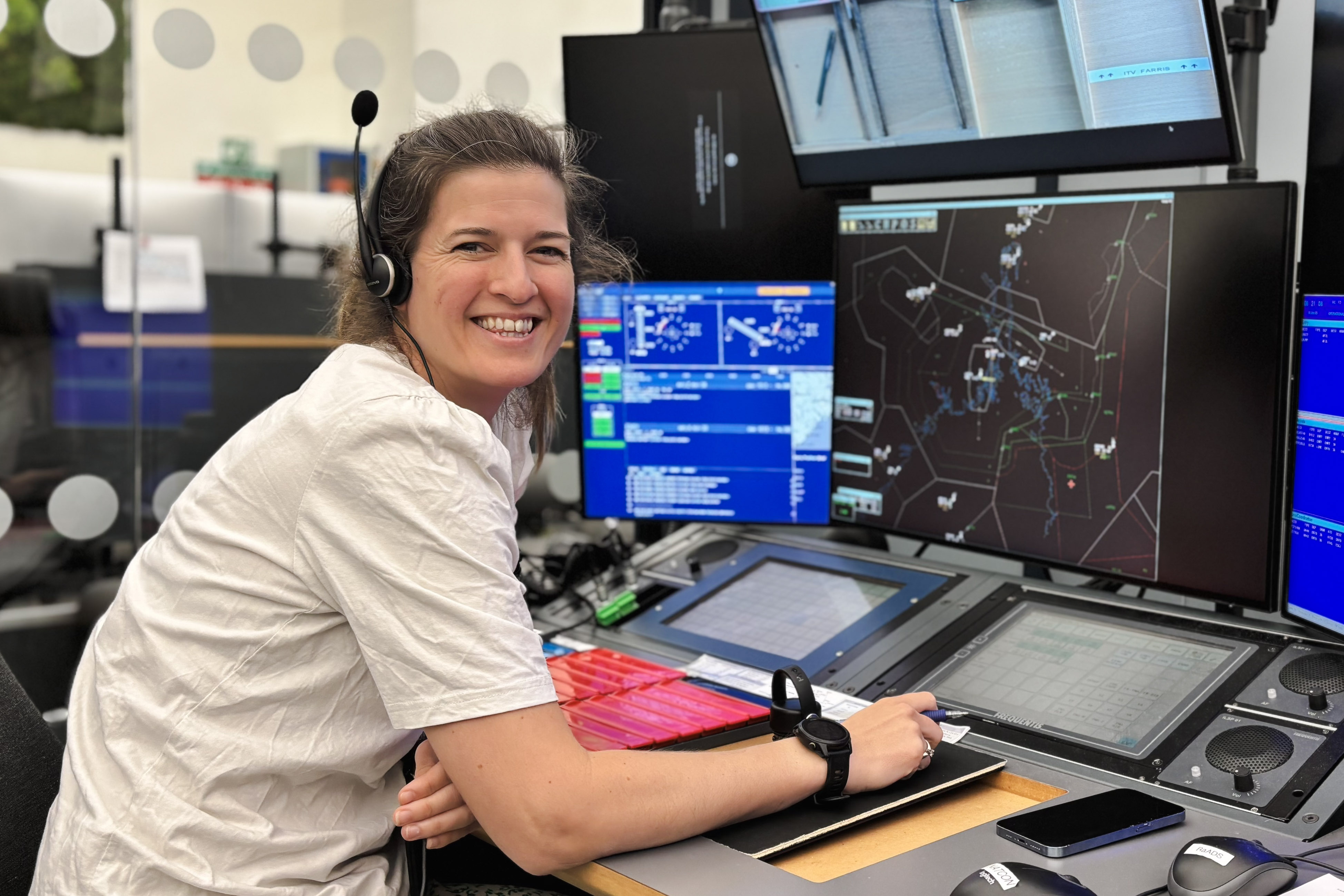 En kvinne i et kontrollrom omgivelse jobber med flere dataskjermer foran seg. Hun smiler til kameraet. Skjermer viser informasjon som kan være relatert til overvåking eller kontrollfunksjoner.