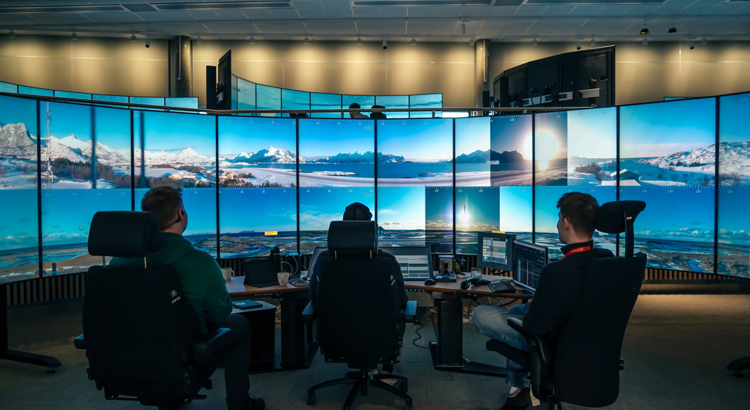 Ein person sit ved eit kontrollrom omgjeve av store skjermar som viser eit landskap av himmel og fjell.