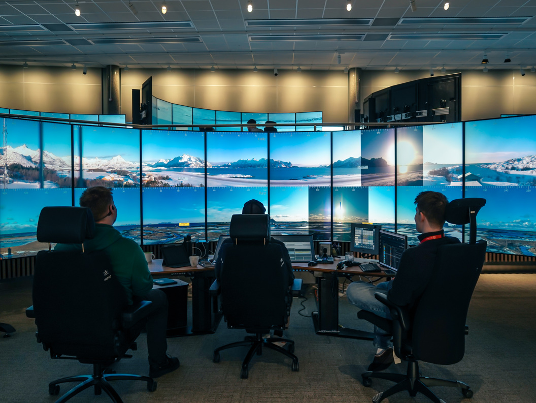 A person sits in a control room surrounded by large screens displaying a landscape of sky and mountains.