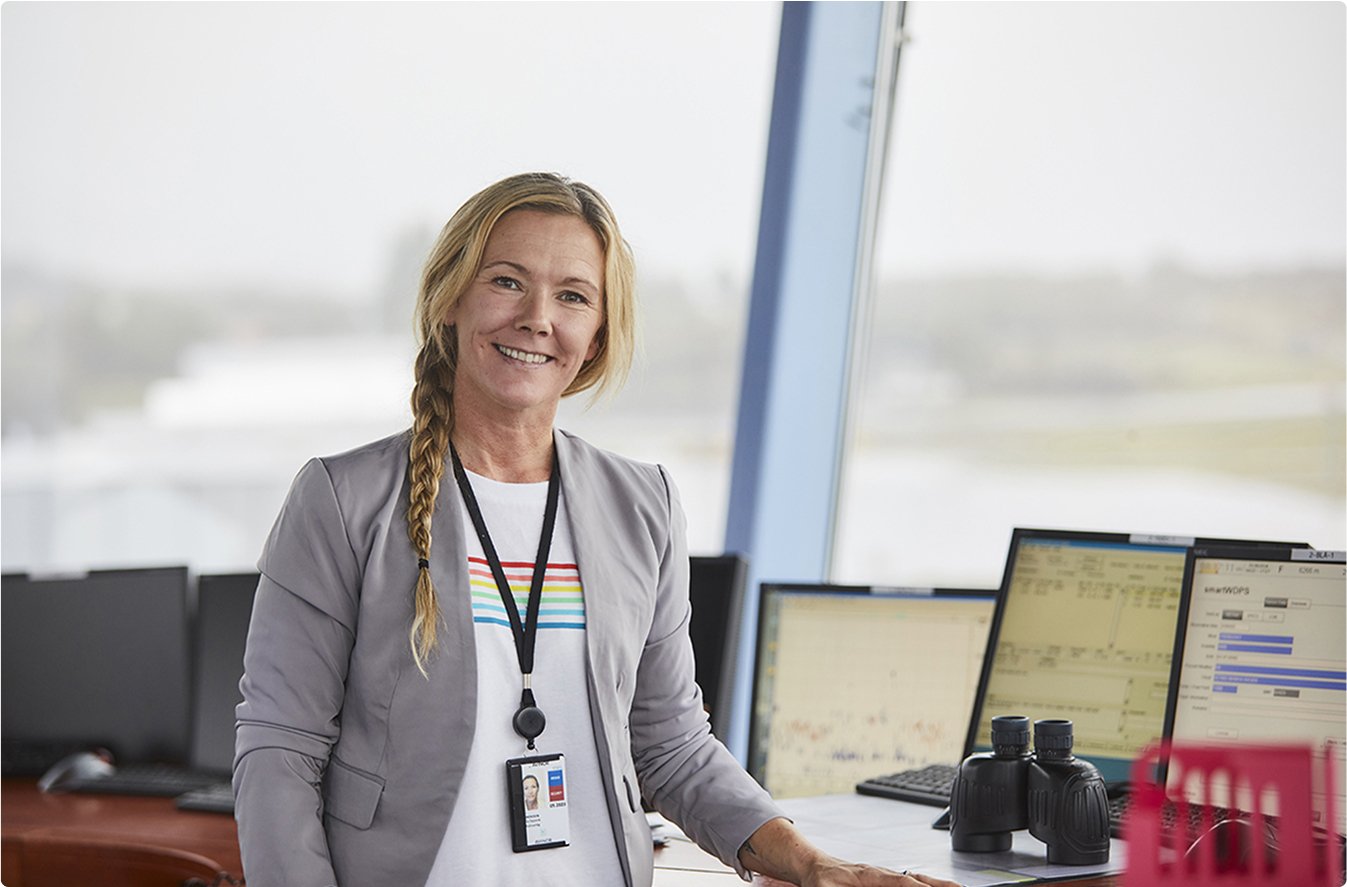 Kvinneleg kontormedarbeidar med oppsett hår og ID-kort står framfor fleire dataskjermar