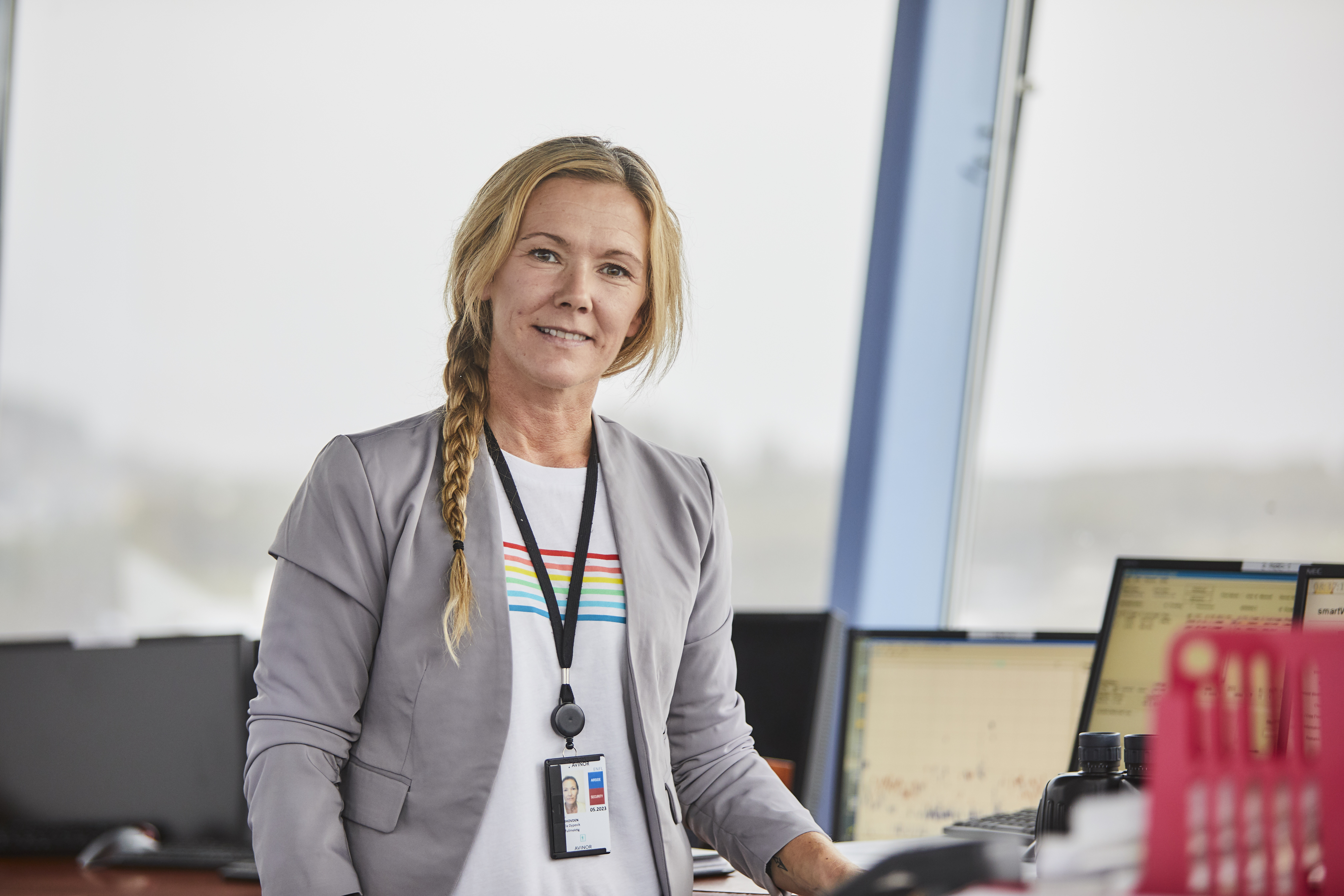 Woman in control tower wearing a gray blazer and ID card, standing in front of screens displaying flight information.