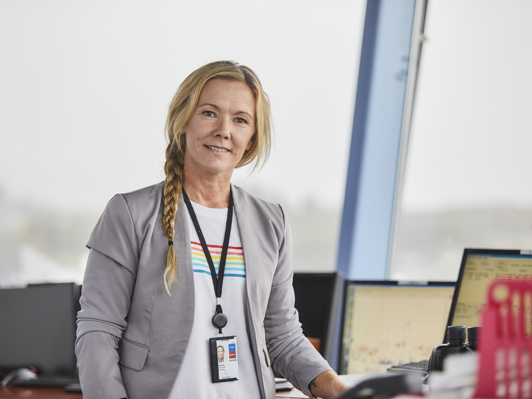 Woman in control tower wearing a gray blazer and ID card, standing in front of screens displaying flight information.