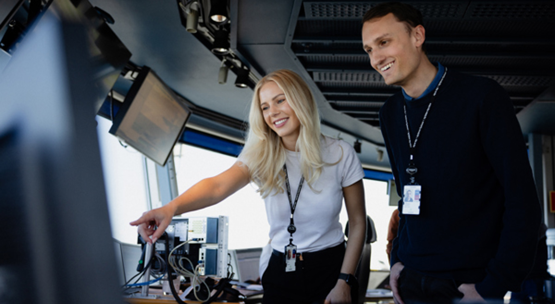 To personar som arbeider i eit moderne kontrollrom, ser på ein skjerm. Den eine peiker mot skjermen med eit smil