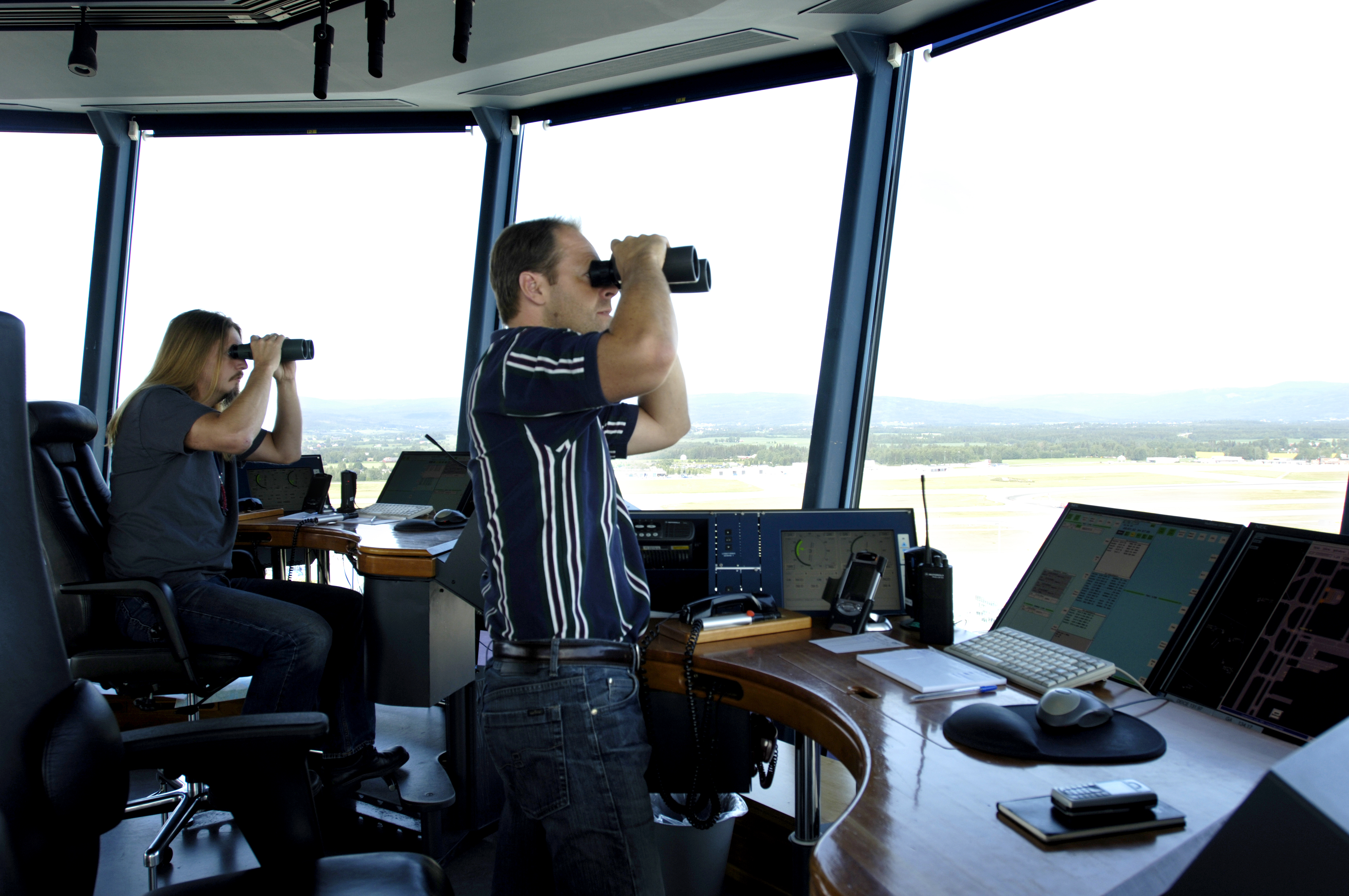 Air traffic controllers in a control tower monitor air traffic using binoculars and computer screens.