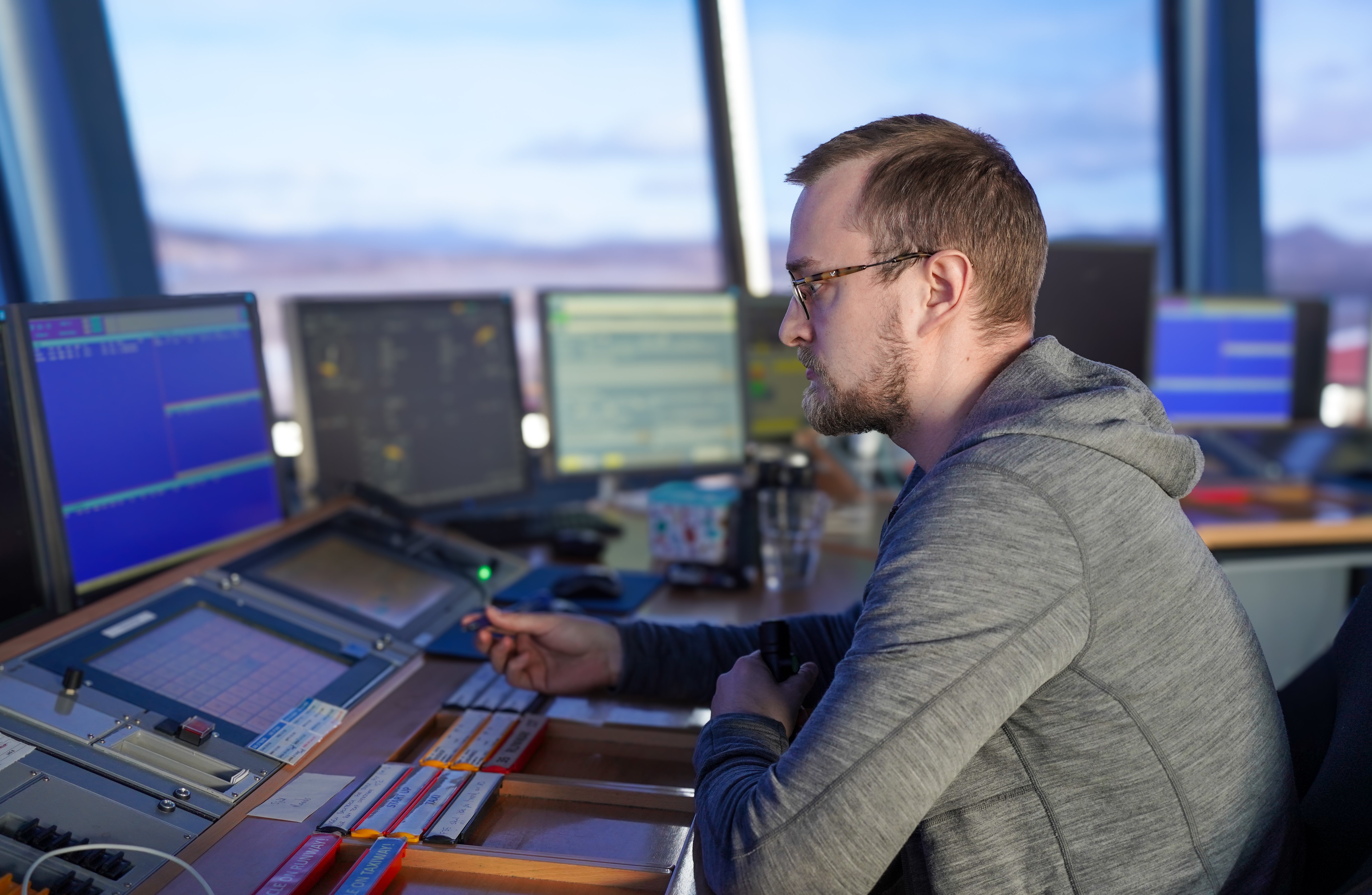 A man in a control room environment is working with several computer screens in front of him. He is wearing a gray hoodie and looks focused. The screens display information that may be related to monitoring or control functions.