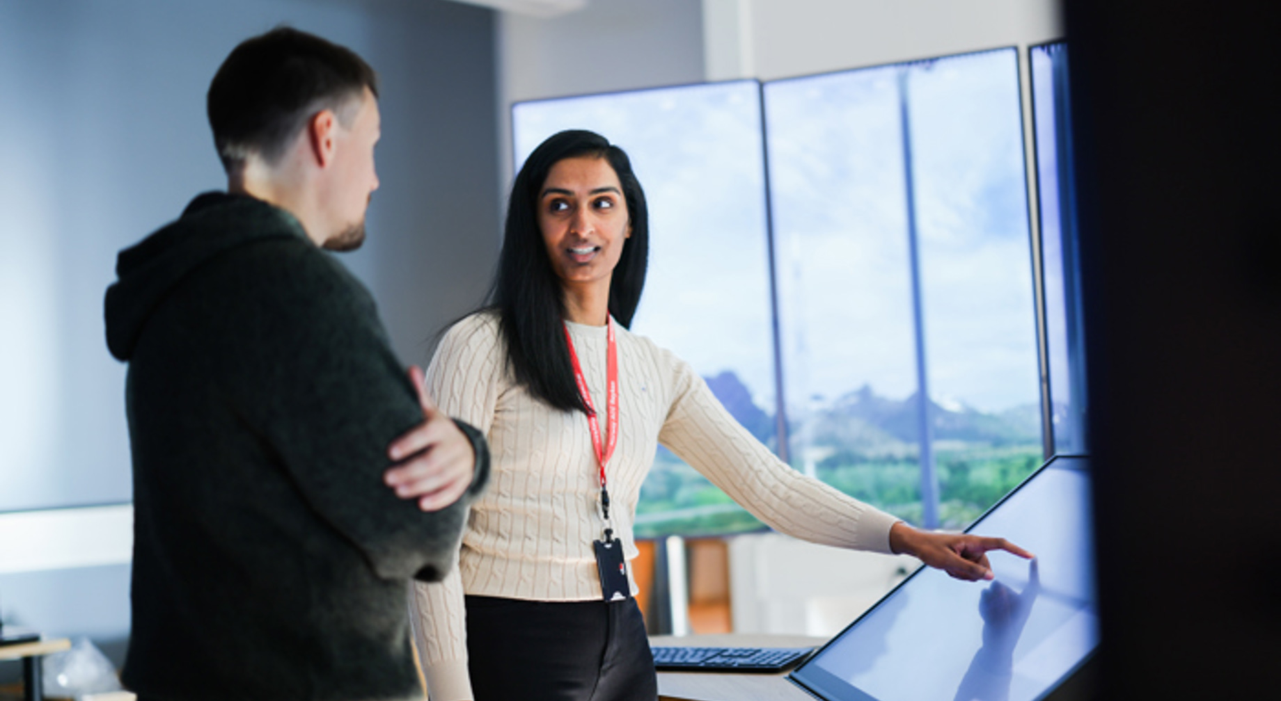 To personer diskuterer foran en stor interaktiv skjerm i et moderne kontormiljø. En kvinne peker på skjermen mens en mann lytter oppmerksomt. Bakgrunn med flere skjermer.