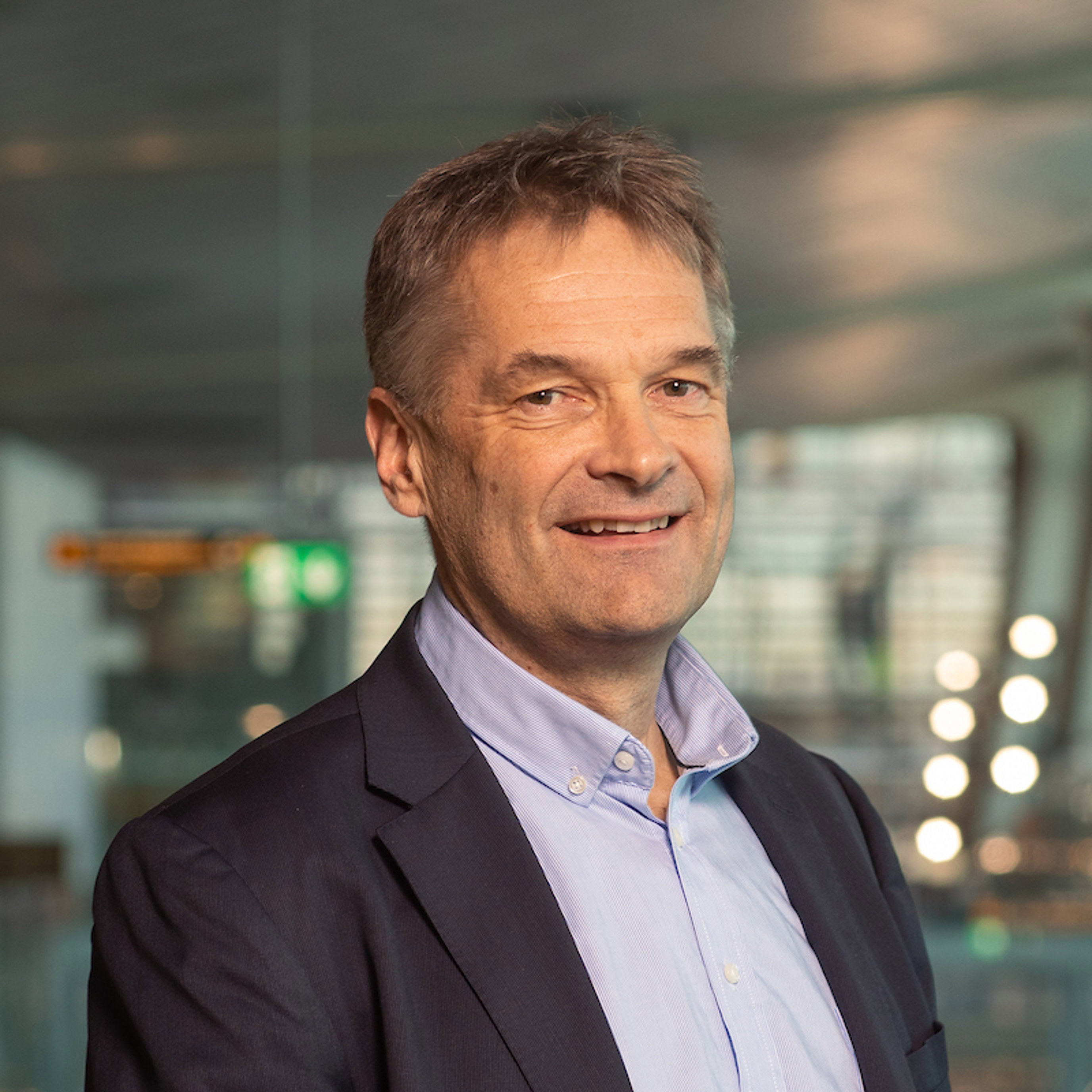 Ein person i dressjakke og skjorte, smilande i eit moderne, profesjonelt innemiljø.