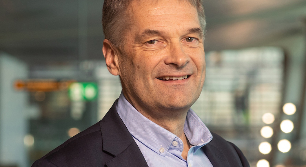 Ein person i dressjakke og skjorte, smilande i eit moderne, profesjonelt innemiljø.