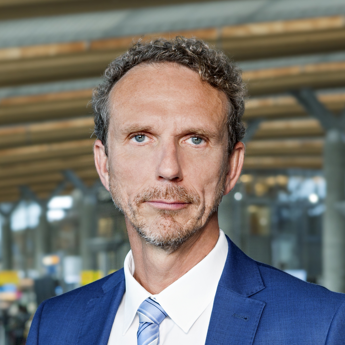 Ein mann i blå dress og slips står innandørs, sett mot ein moderne, romsleg bakgrunn med trepaneltak.