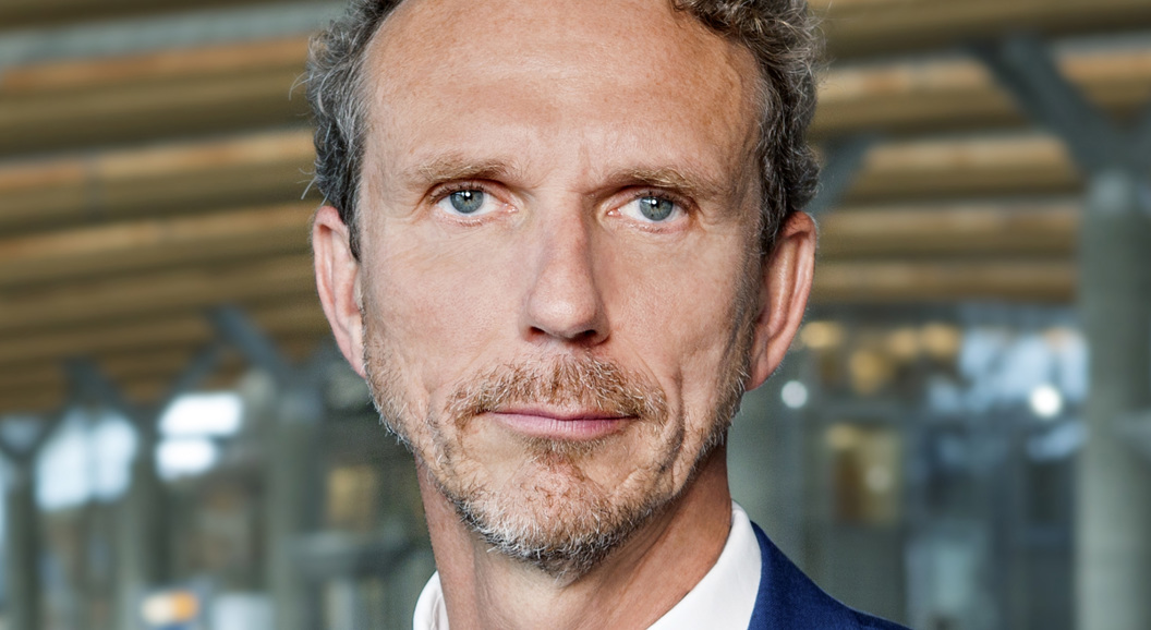 Ein mann i blå dress og slips står innandørs, sett mot ein moderne, romsleg bakgrunn med trepaneltak.