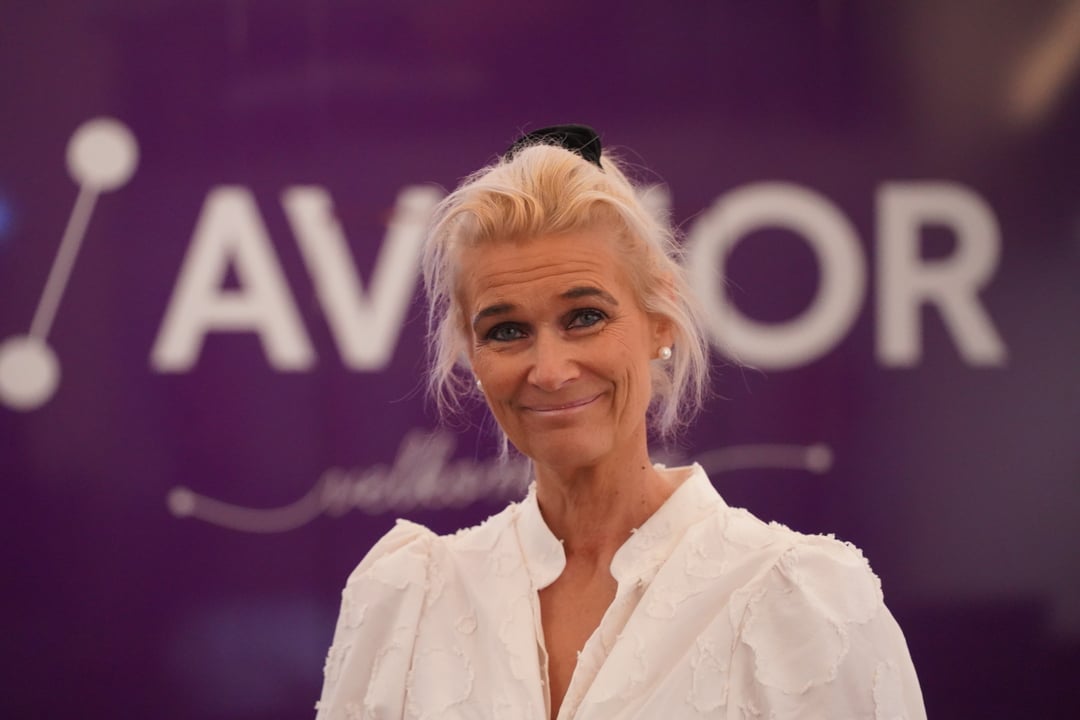 Kvinne i hvit kjole smiler foran en lilla bakgrunn med utydelig tekst.