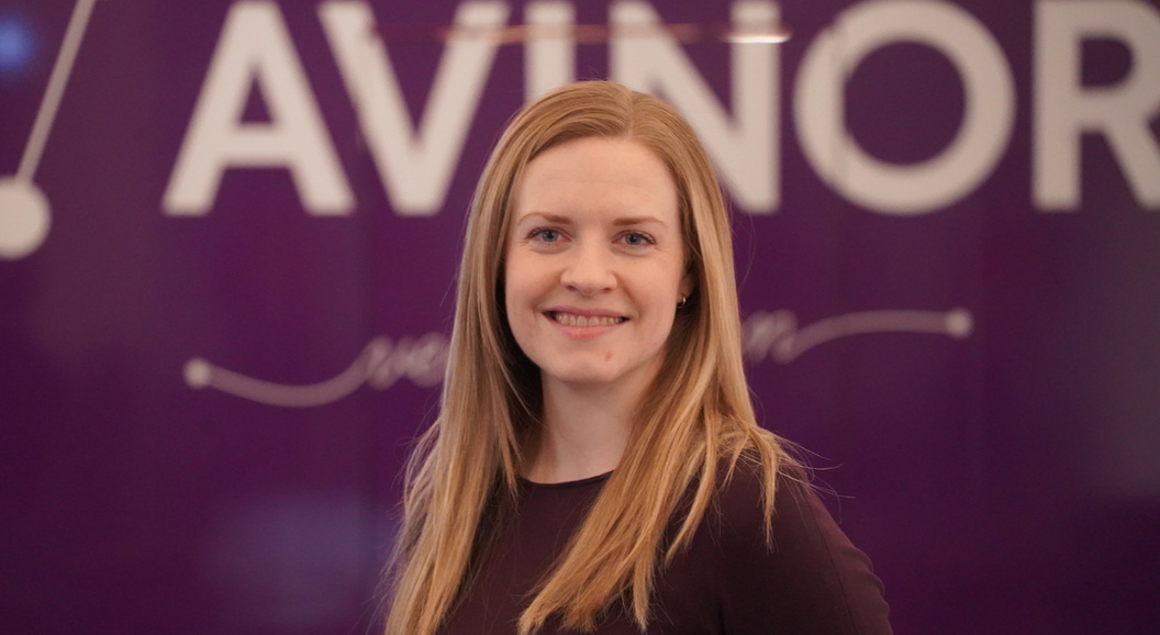 Kvinne med langt blondt hår smiler framfor ein lilla bakgrunn med teksten "AVINOR".