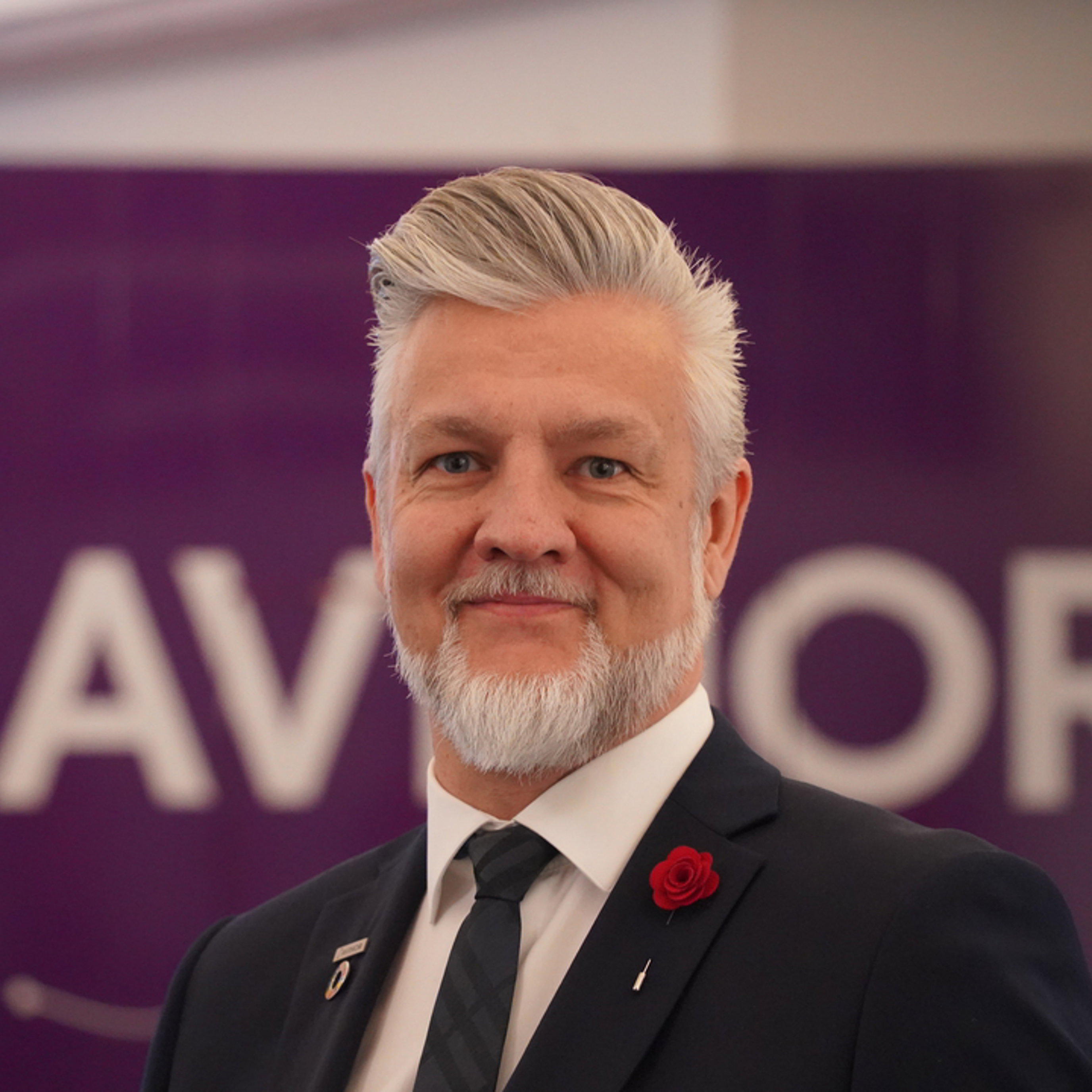 Forretningsmann med grått hår, iført mørk dress og rød rosepin, foran et banner som delvis viser bokstaver.