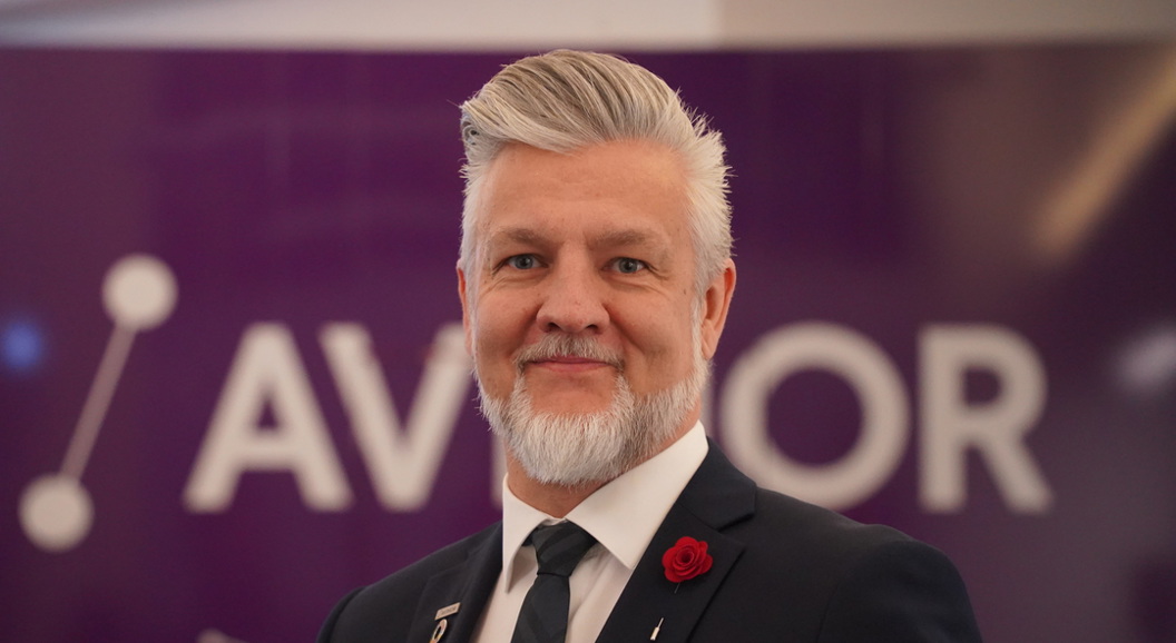 Forretningsmann med grått hår, iført mørk dress og raud rosepin, foran eit banner som delvis viser bokstavar.