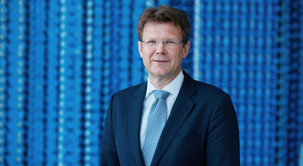 Ein mann i dress og slips står framfor ein blå bakgrunn med mønster.