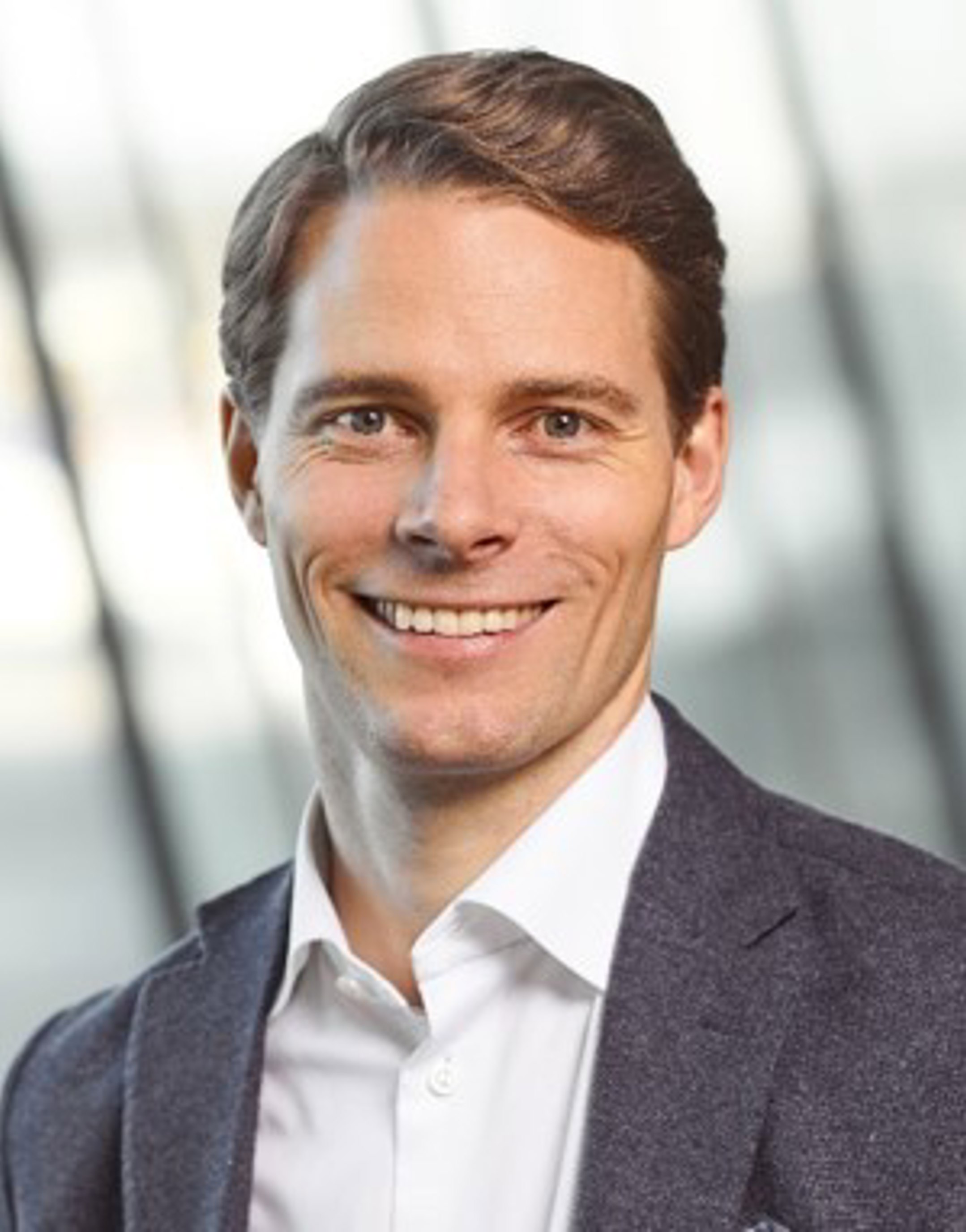 Mann i forretningsantrekk med kvit skjorte og mørkeblå blazer smiler mot kameraet. Bakgrunnen er uskarp
