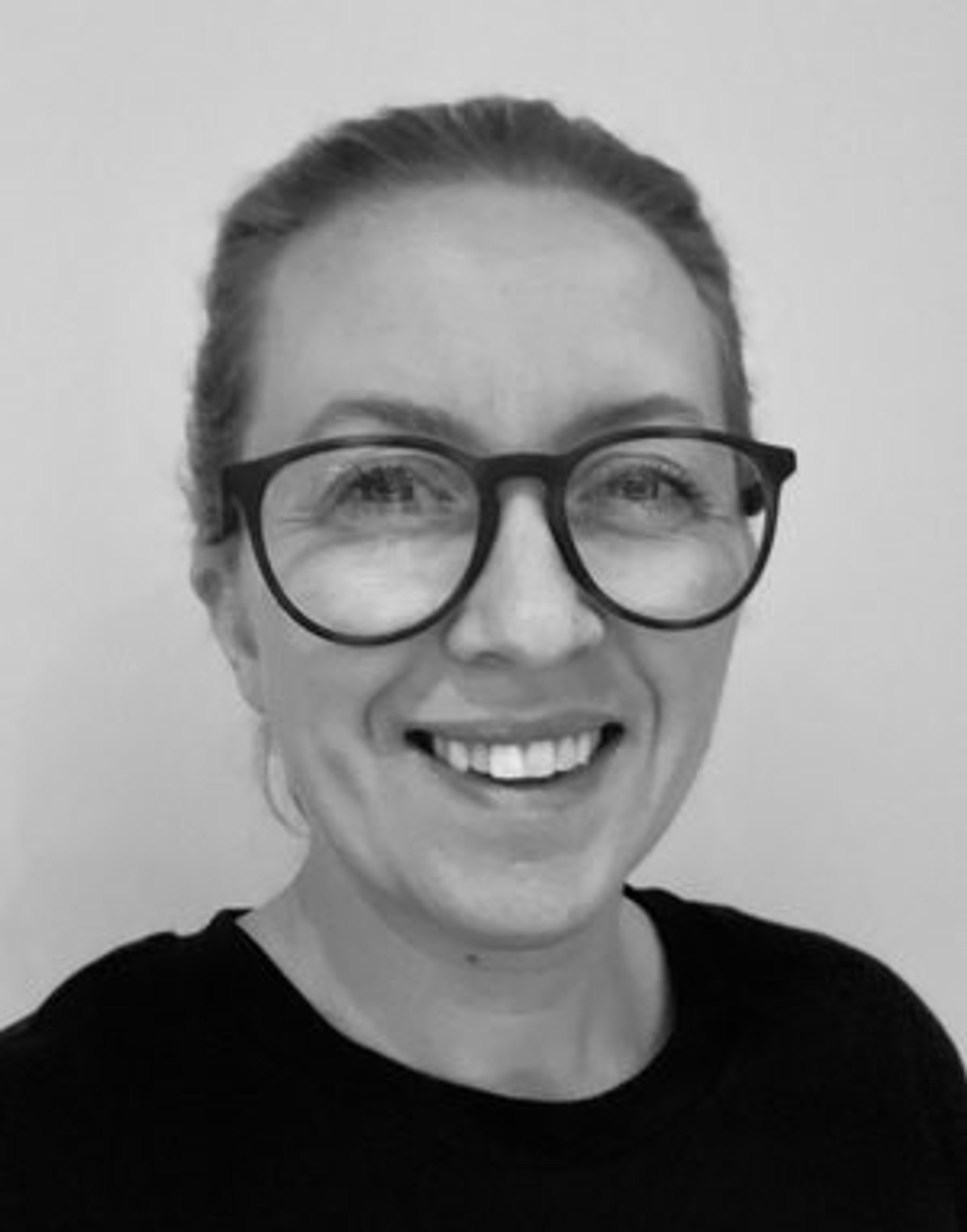 Kvinne med briller og smil, kledd i svart genser, mot ein nøytral bakgrunn