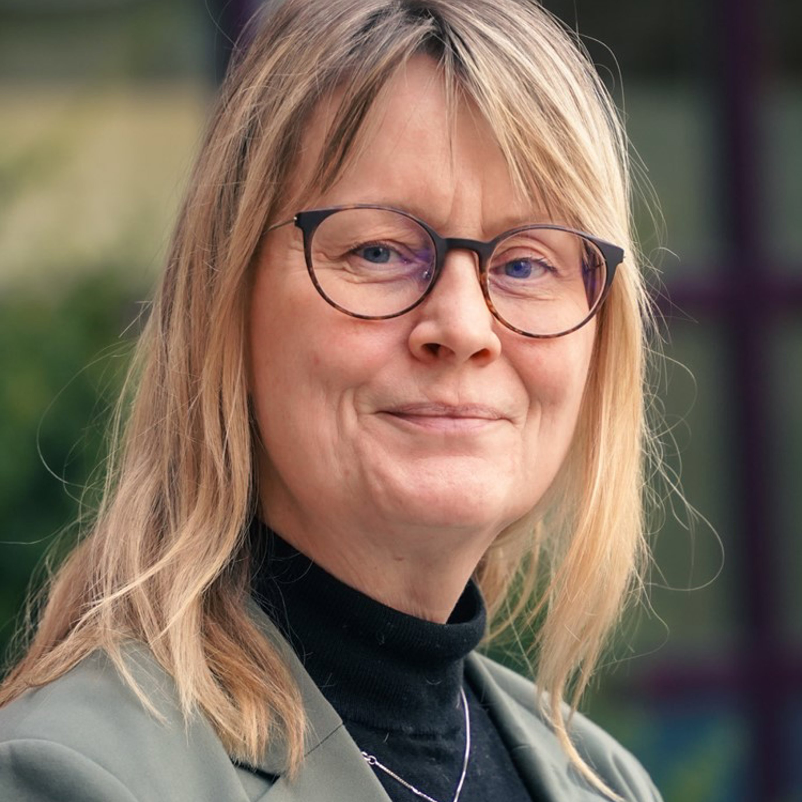 Smilende kvinne med briller og blondt hår, iført en grå jakke og en svart høyhalset genser, stående utendørs