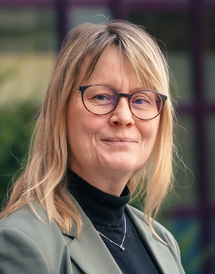 Smiling woman with glasses and blonde hair, wearing a gray jacket and a black turtleneck sweater, standing outdoors