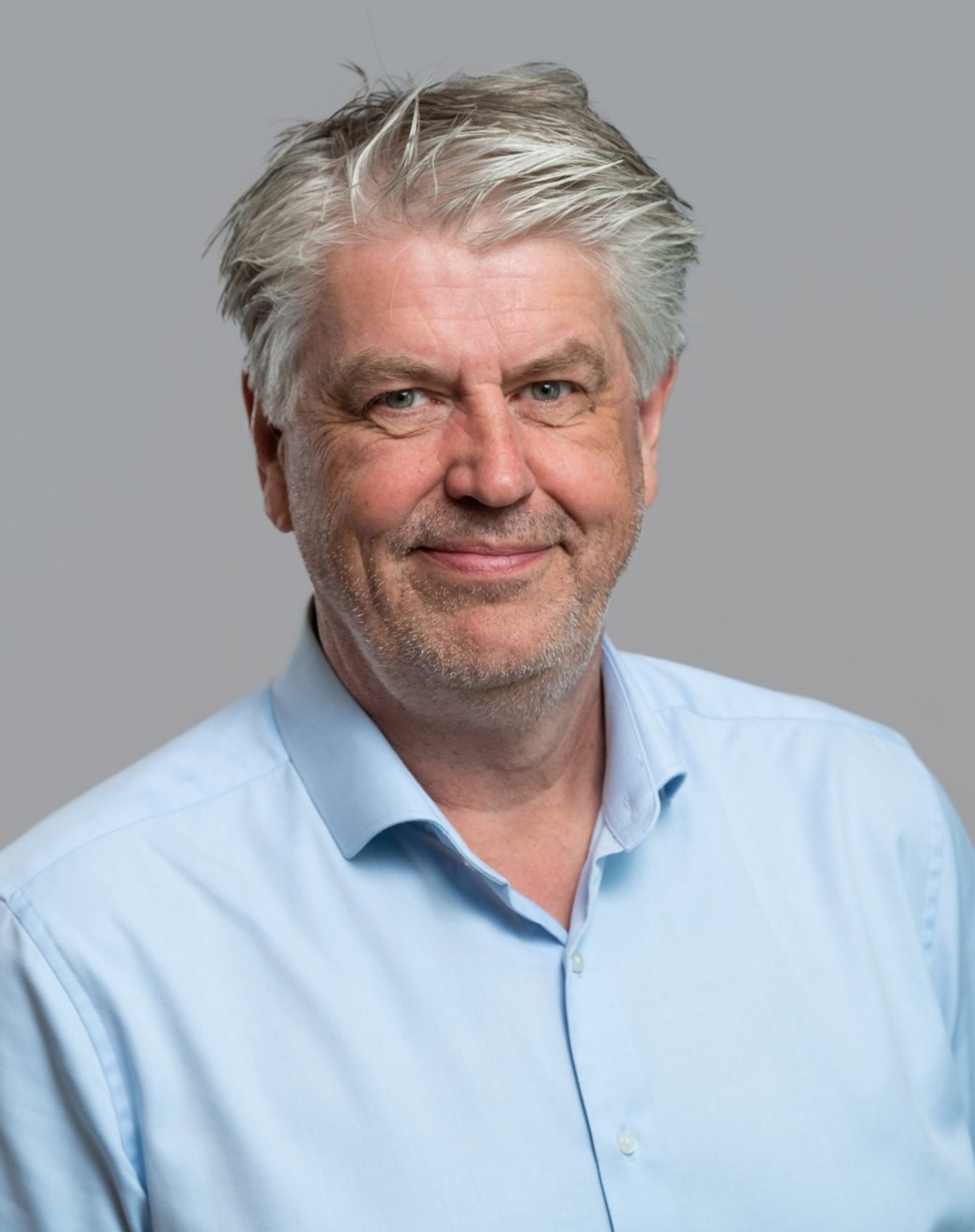 Eldre mann med grått hår, iført lyseblå skjorte, smiler foran grå bakgrunn