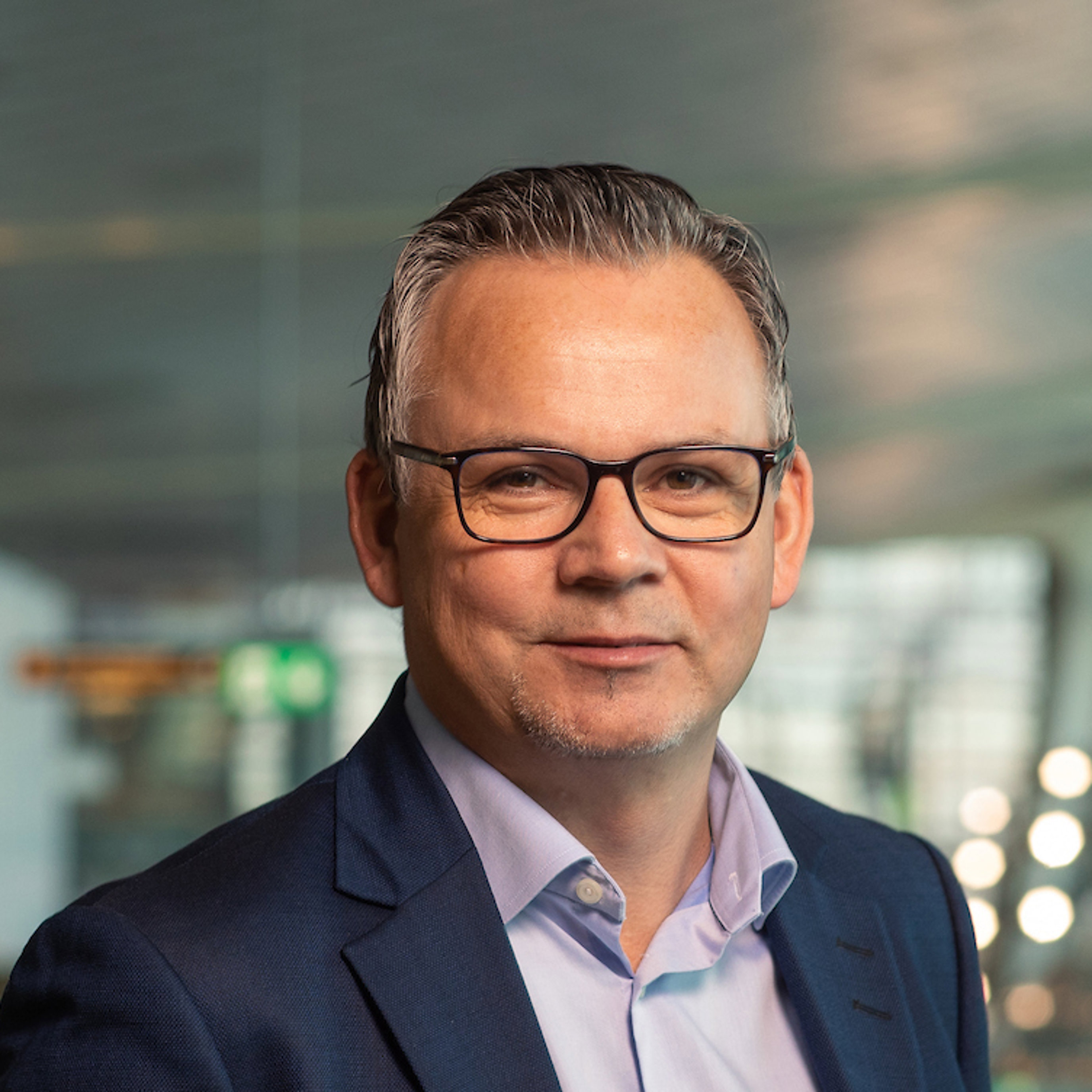 En smilende mann med briller og en blå dressjakke i en moderne kontoromgivelse.