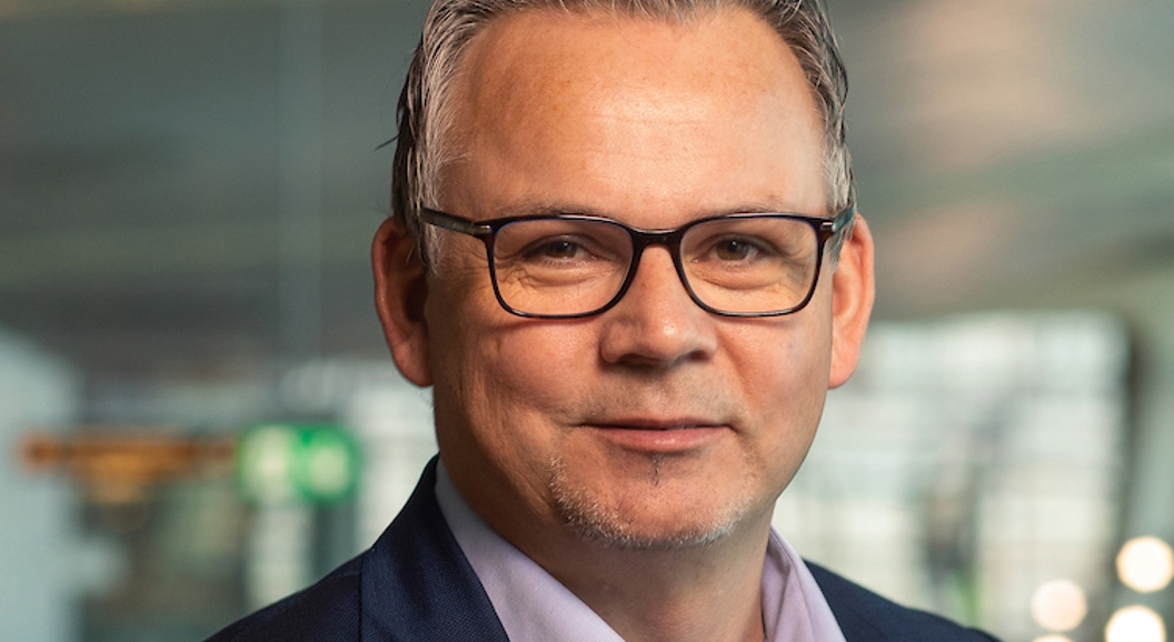 Ein smilande mann med briller og ei blå dressjakke i ein moderne kontoromgivnad.