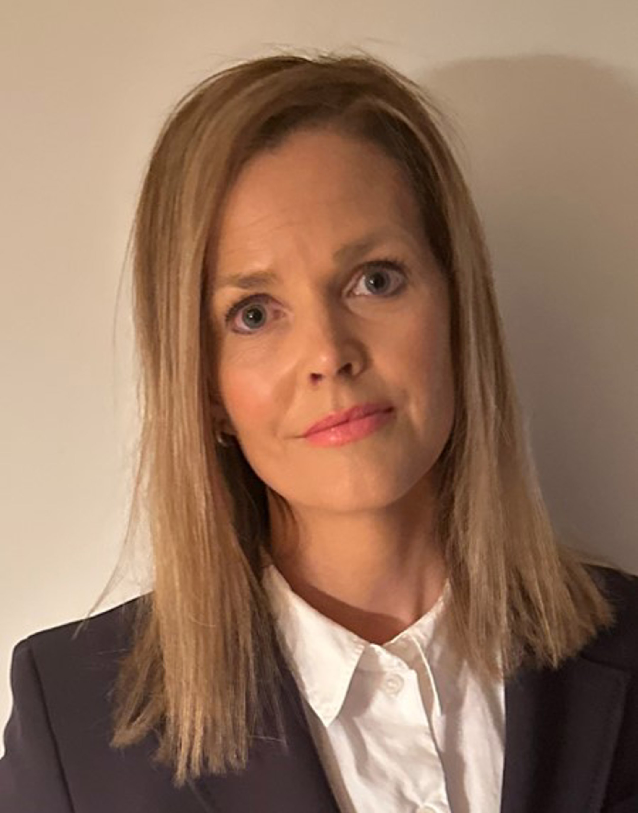 Kvinne med blondt, skulderlangt hår i svart blazerjakke og kvit skjorte, ser direkte mot kameraet, nøytral bakgrunn