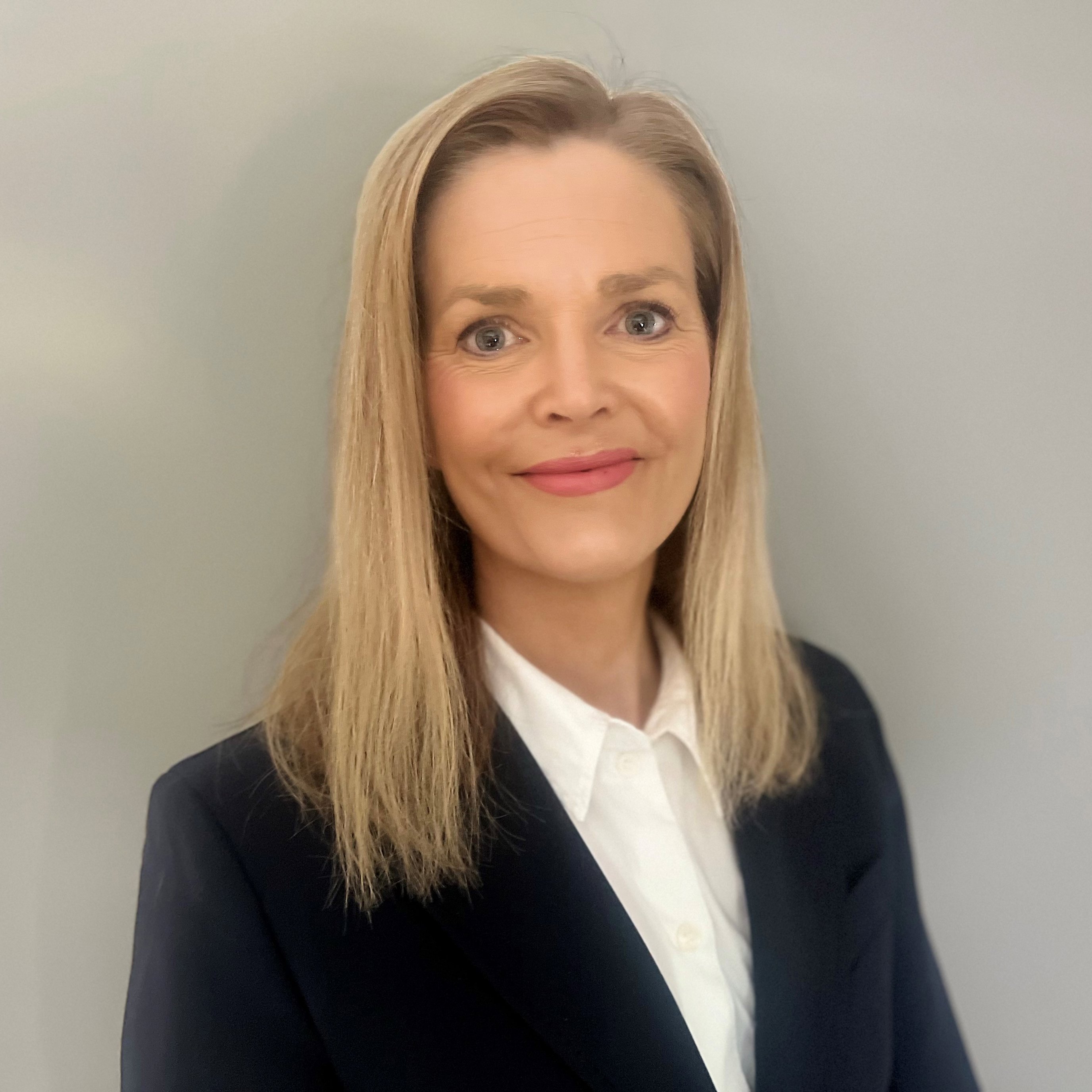 Kvinne med langt blondt hår iført en mørk dressjakke og hvit skjorte, foran en grå bakgrunn.